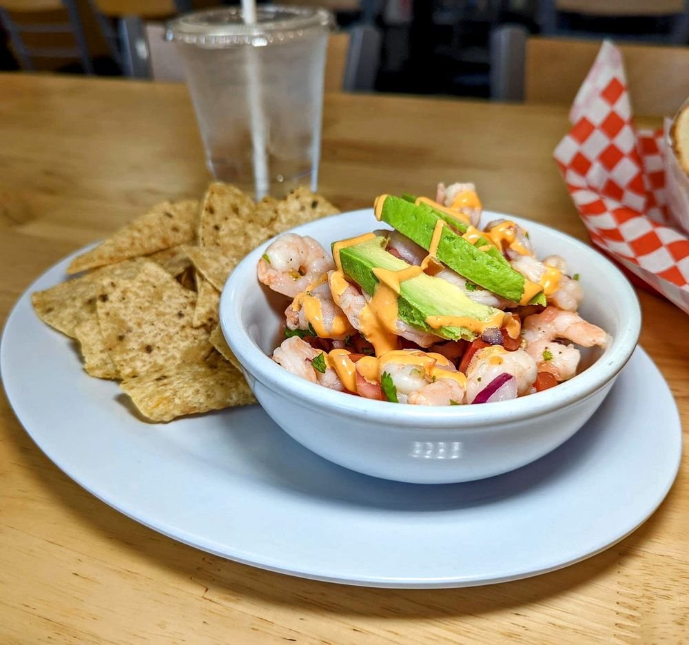 Shrimp ceviche with avocado and diced vegetables served with tortilla chips, cold drink in a plastic cup and a basket of food in the background