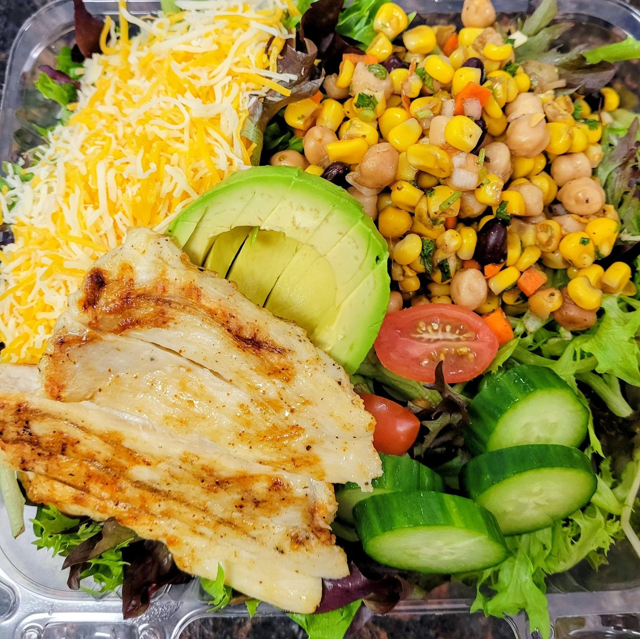 Mixed salad with shredded cheese, chickpeas, corn, cherry tomato, cucumber slices, avocado, grilled chicken, and leafy greens.