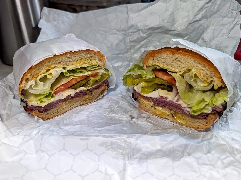 A cut sandwich with lettuce, tomato, pickles, mayonnaise, and sliced roast beef on a bun, wrapped in white paper.