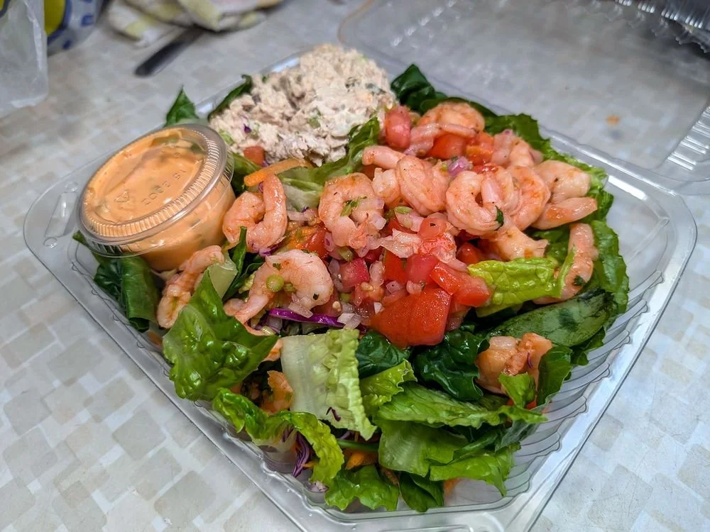 Lettuce salad with cooked shrimp, chopped tomatoes, diced onions, a scoop of seafood salad, and a small container of salad dressing.