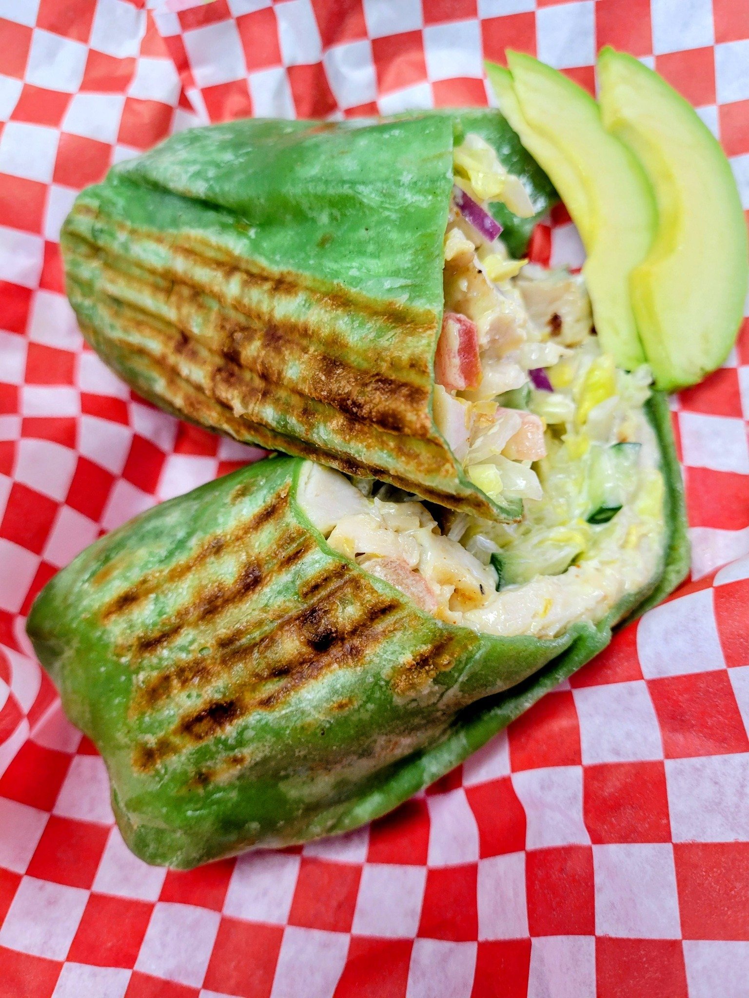 Grilled chicken wrap with lettuce, tomato, and sauce, sliced in half with avocado slices on the side, on red and white checkered paper.