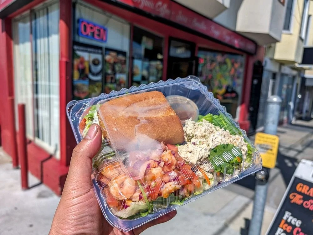 Container with bread, shrimp salad, lettuce, side of potato salad, and a small cup of dressing, held in front of a storefront with an 'OPEN' sign.