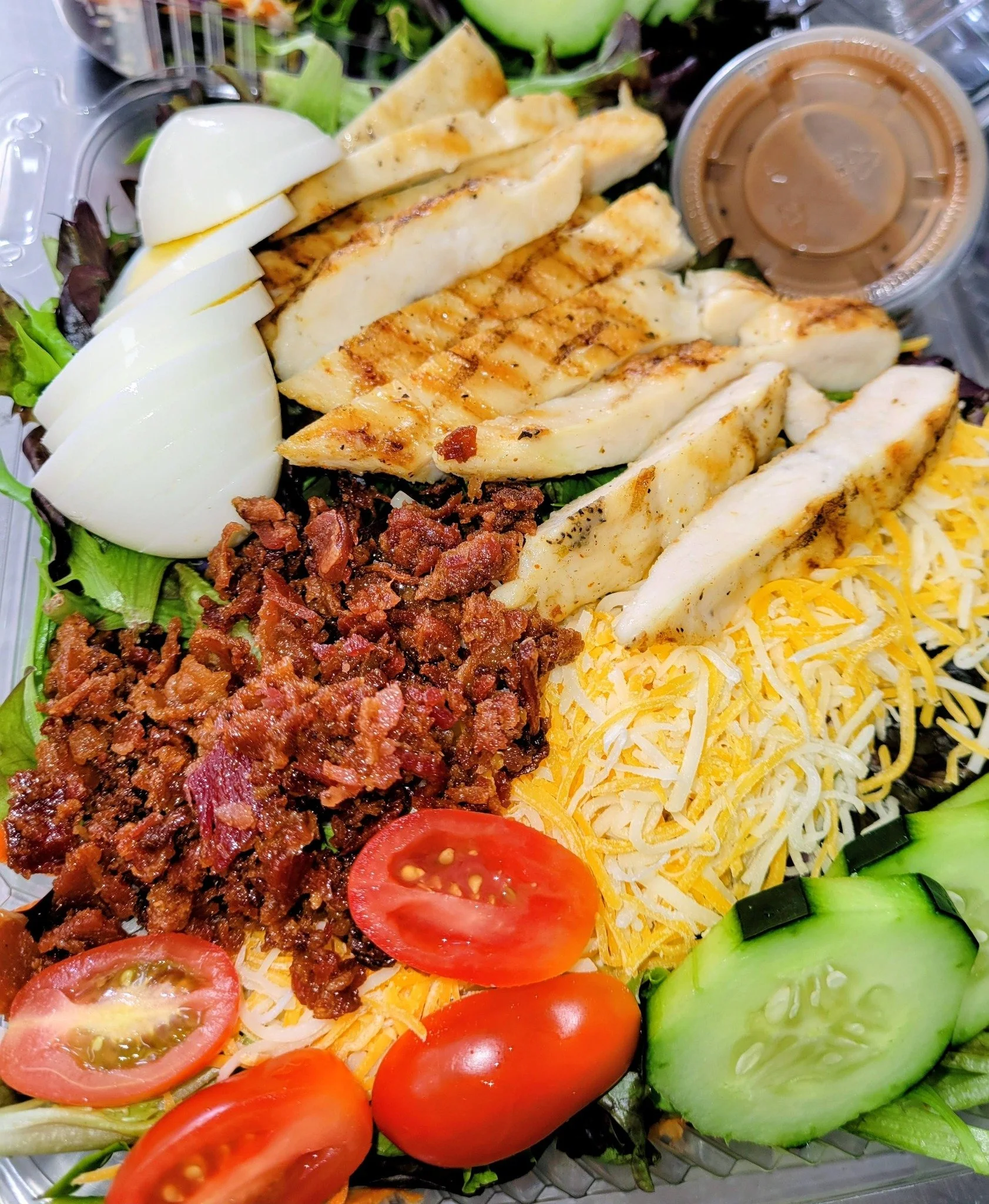 Assorted salad with grilled chicken strips, hard-boiled eggs, cherry tomatoes, shredded cheese, ground beef, cucumber slices, and a small container of dressing on a bed of mixed greens.