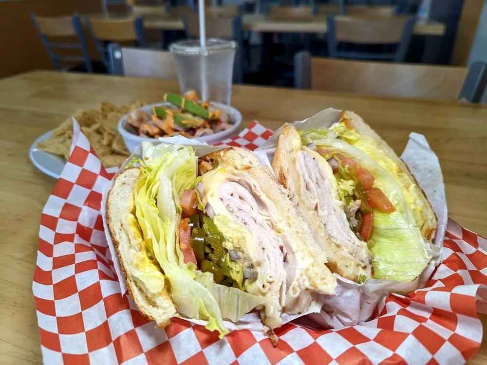 Club sandwich with lettuce, tomato, pickles, and turkey on toasted bread, served with chips, in a casual restaurant setting.