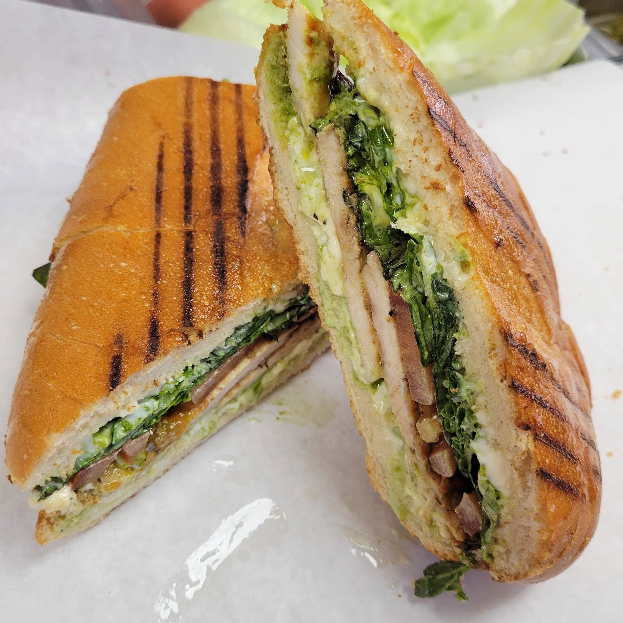Grilled sandwich with lettuce, tomato, and possibly chicken or turkey, cut in half and served on a white surface with a side of lettuce.