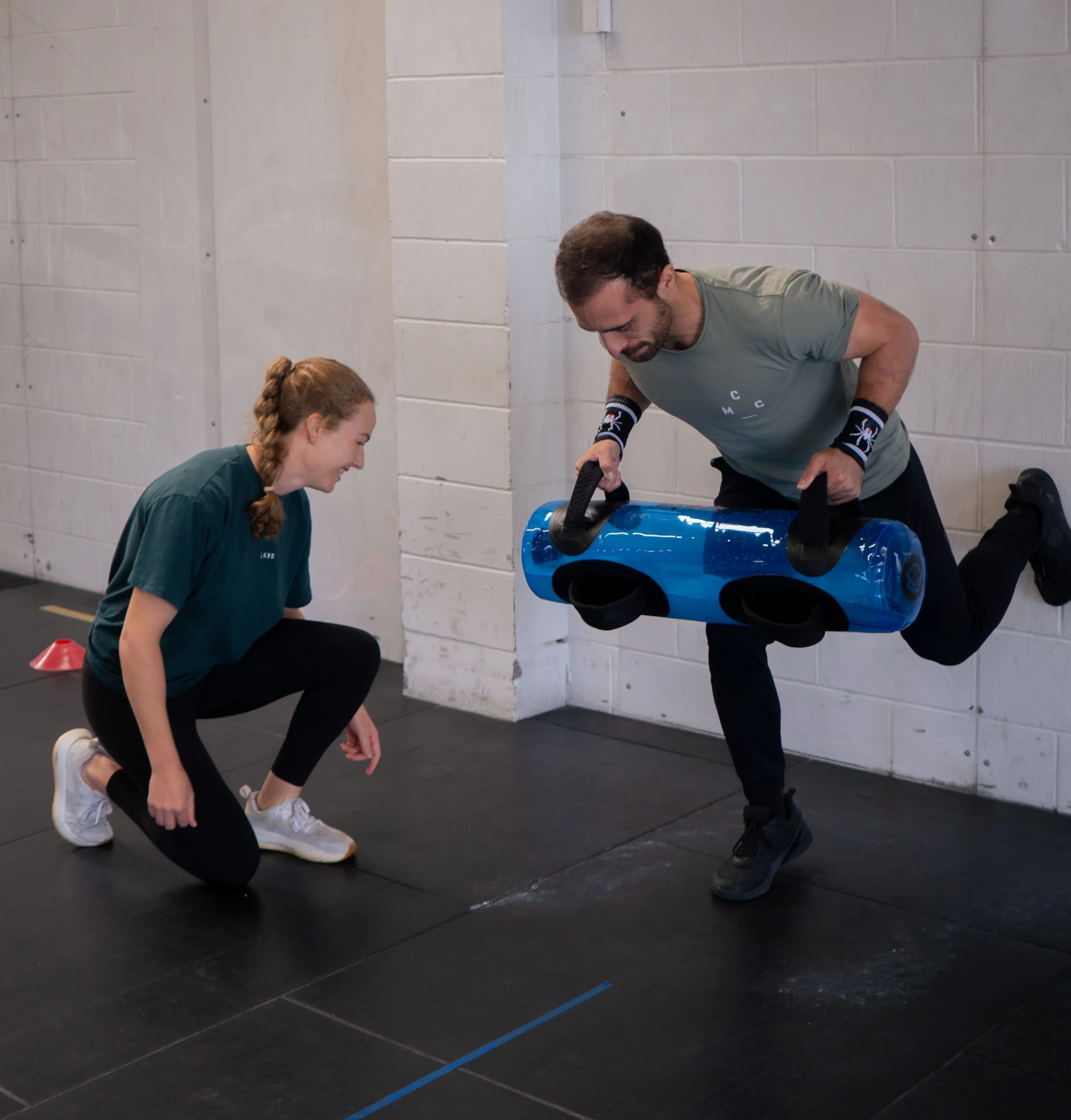 A woman kneeling on the floor and a man standing next to her, both smiling and working together in a gym or fitness area with black rubber flooring and white brick walls.
