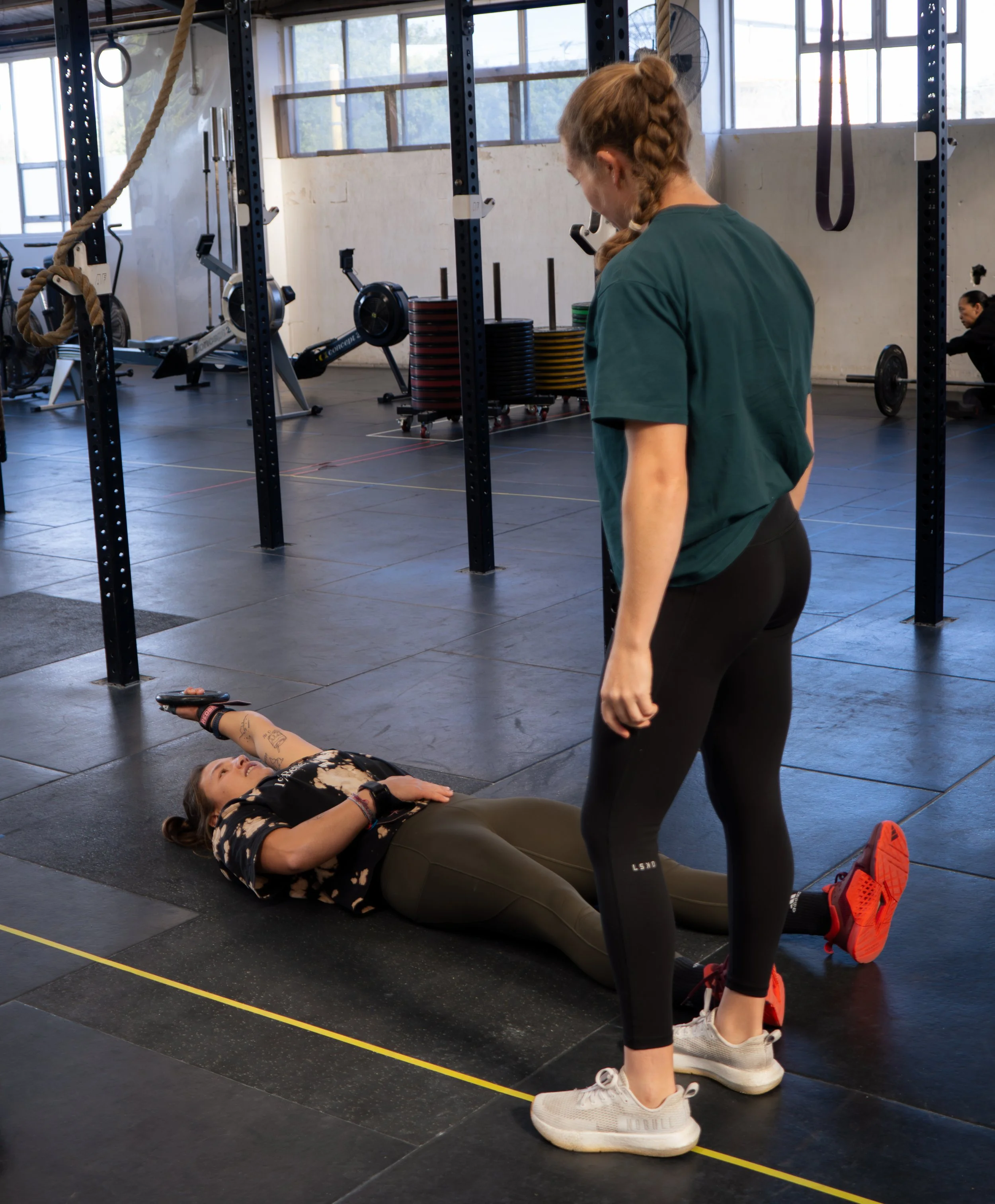 A woman lying on her back on a gym floor with a woman standing nearby, in a gym with workout equipment in the background.