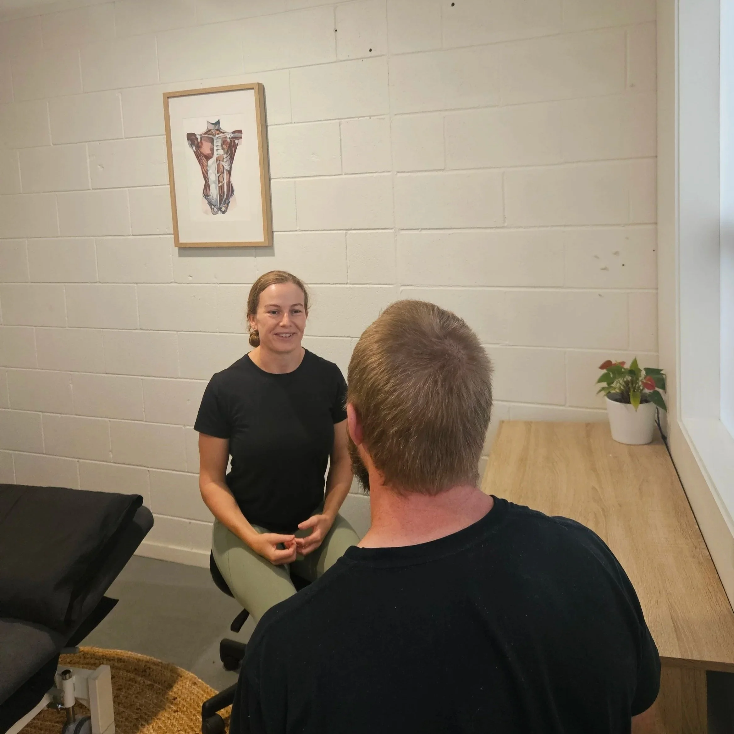 A woman with glasses and braided hair smiling, seated at a light-colored table in a room with white brick walls and framed artwork, with a person in black in the foreground and a potted plant on the side.