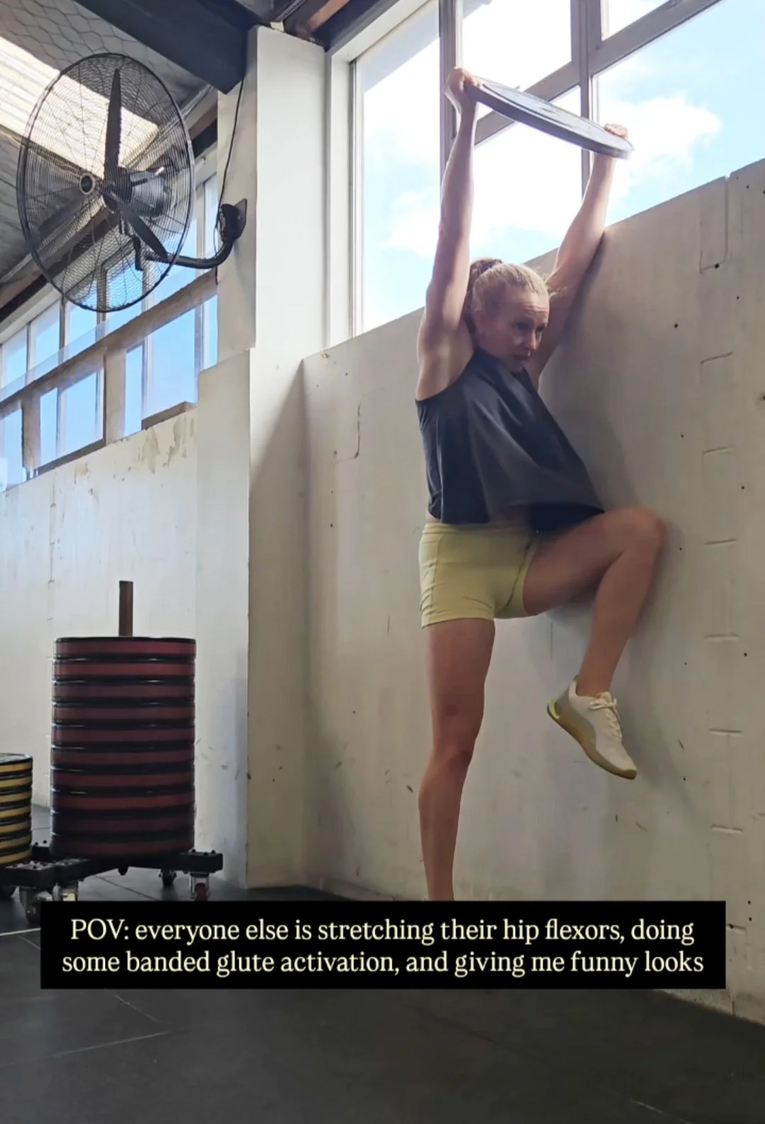 A woman is stretching her hip flexors while standing on one leg with her other leg placed against a wall. She is holding a weight above her head, wearing a black tank top, yellow shorts, and sneakers in a gym with large windows and a fan. There are weight plates stacked on a rack nearby.