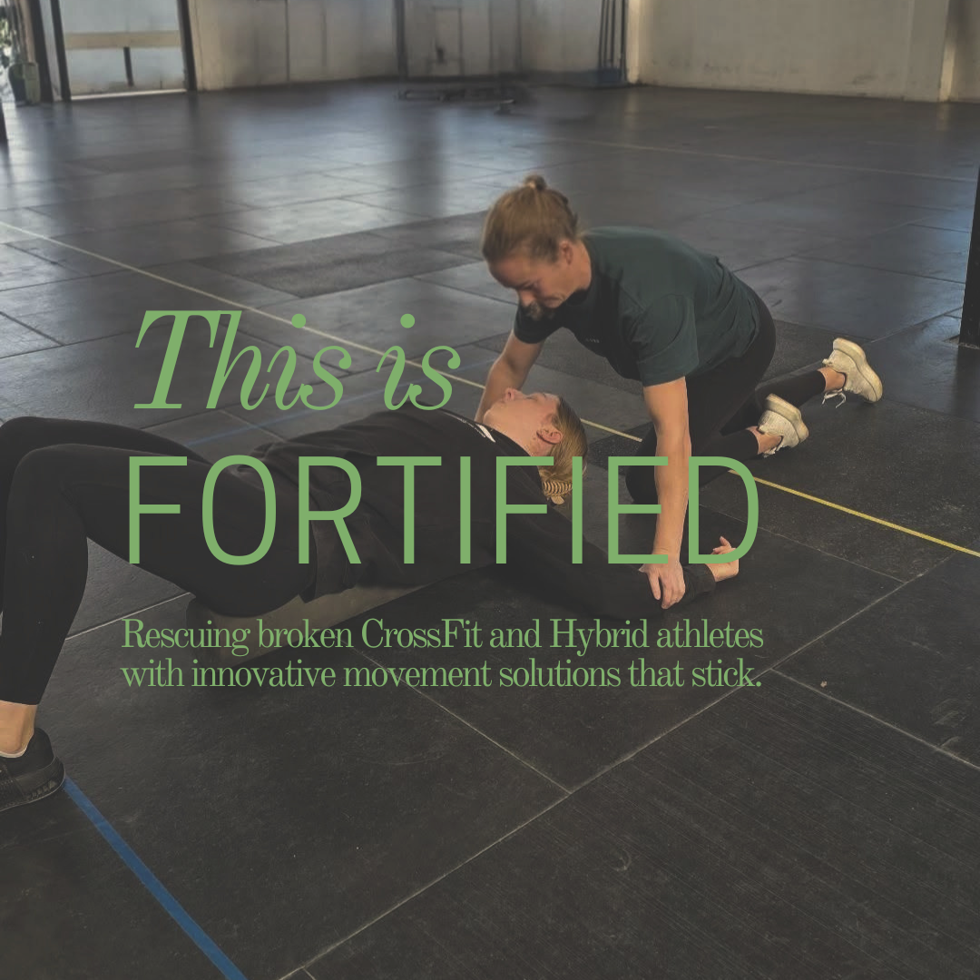 A woman lying on the floor performing an exercise while another woman, kneeling, assists her in a gym with a black rubber floor and gray walls.