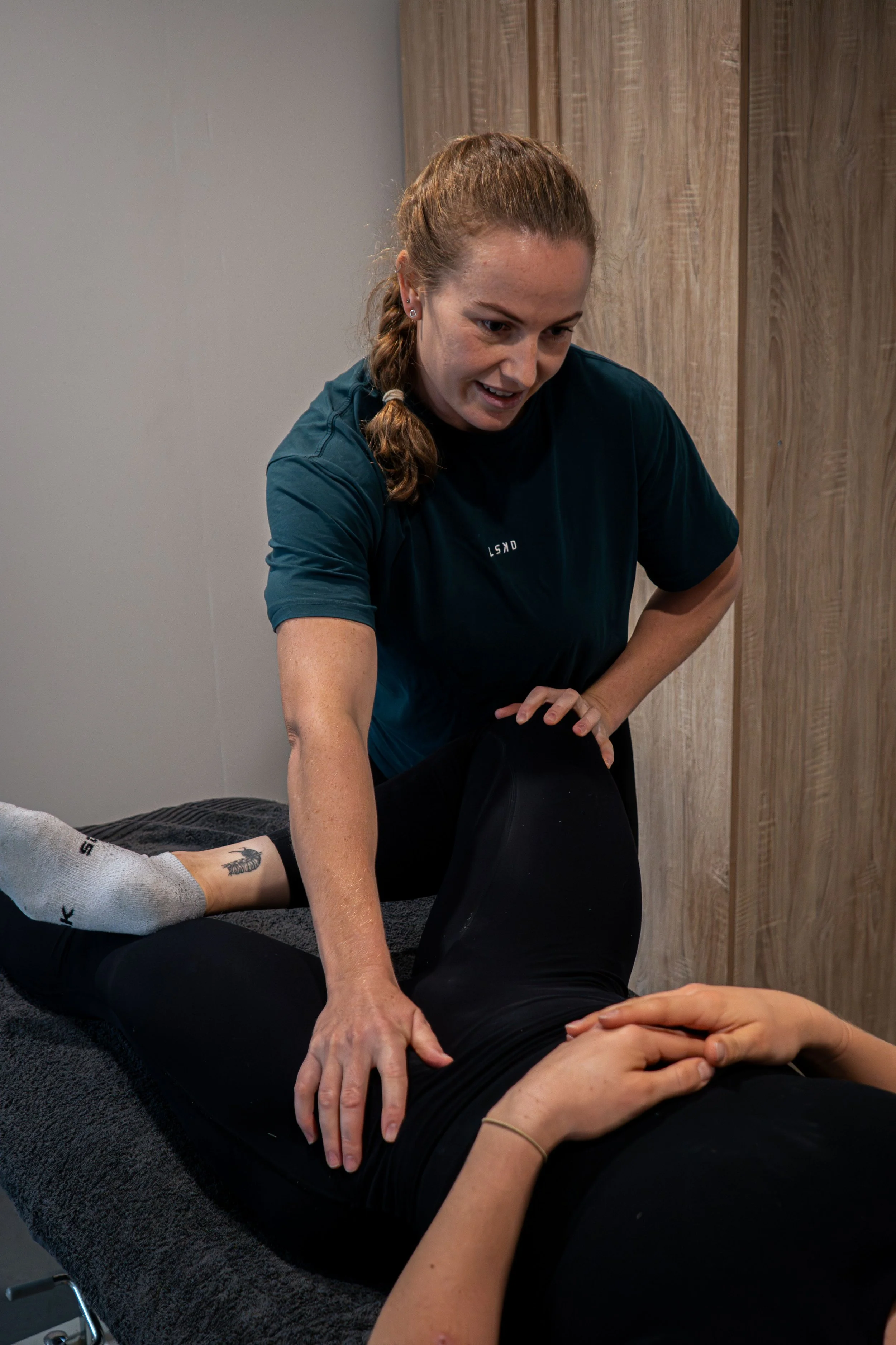 A woman undergoing physical therapy, lying on a treatment table while a female therapist supports her leg during a stretching or mobility exercise.
