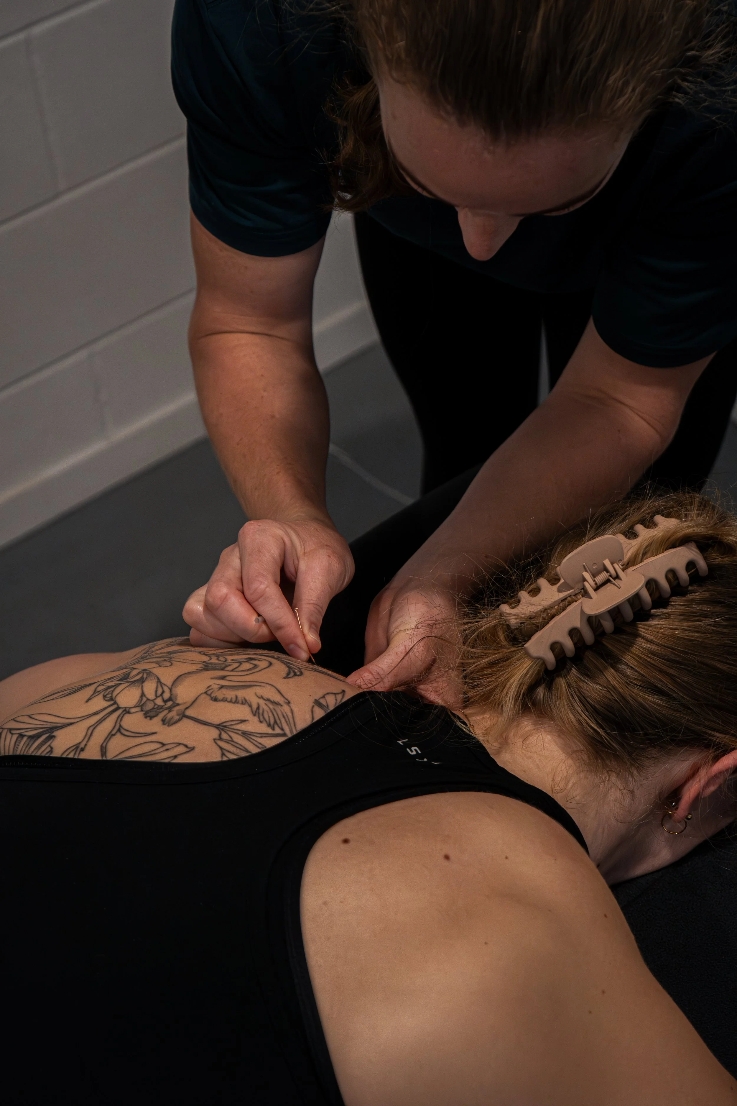 A person receiving CPR training, lying on their back, with a trainer performing chest compressions on them. The person has tattoos on their chest and is wearing a black top. The trainer is leaning over, focused on the procedure.