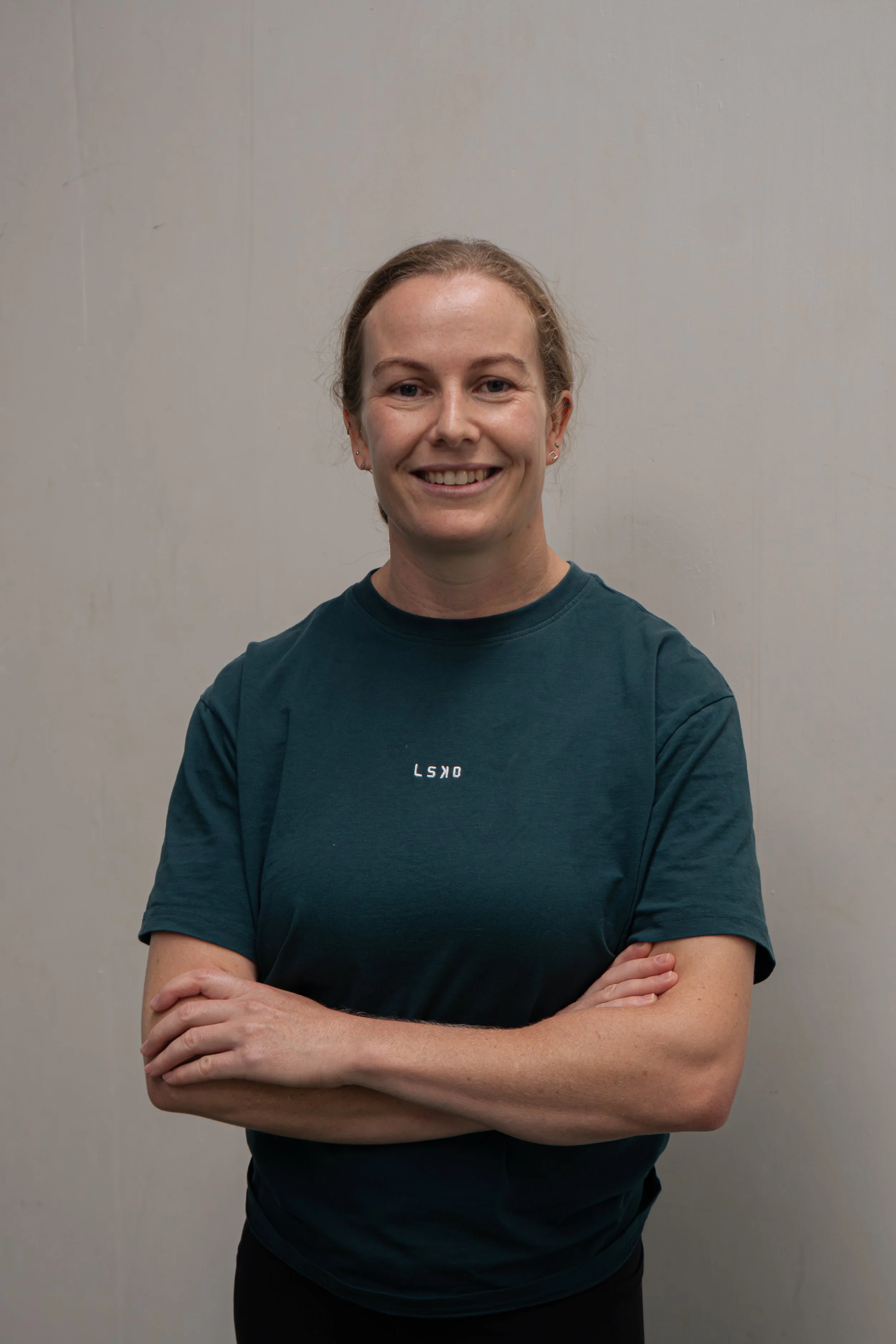 A woman standing with arms crossed in front of a beige wall, smiling, wearing a dark teal T-shirt with small white text, and has brown hair pulled back.