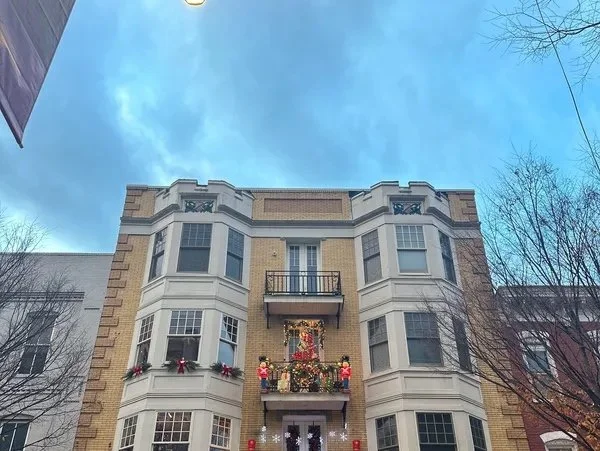 A four-story apartment building decorated for Christmas with wreaths and hanging ornaments, with a small balcony on the second floor and large windows on each floor, under a blue sky with a few clouds and leafless trees on either side.