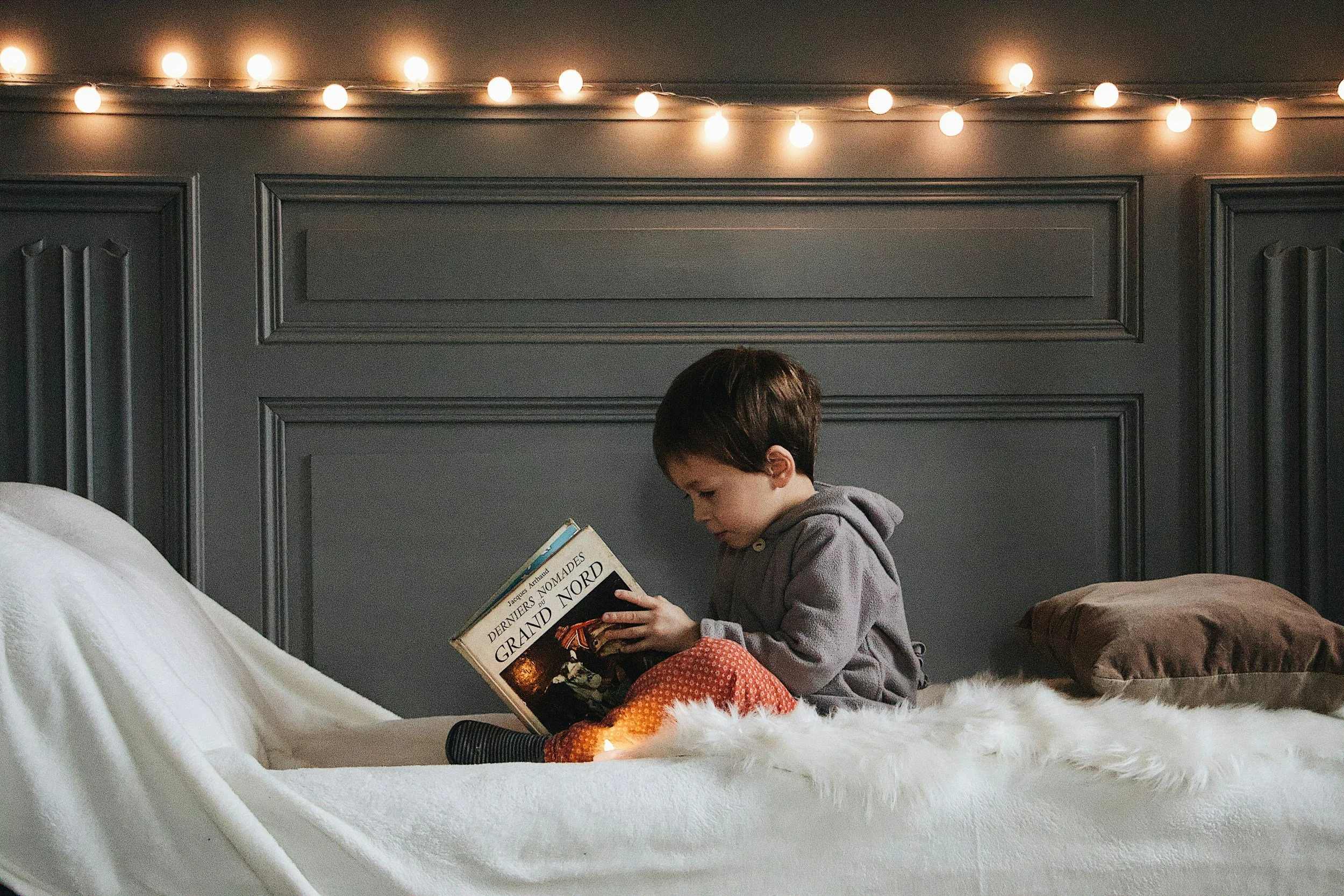 A young boy sitting on a bed with white bedding and a furry throw, reading a book titled "Les Derniers Noms du Grand Nord" in a room with dark gray paneled walls and string lights hanging across the ceiling.