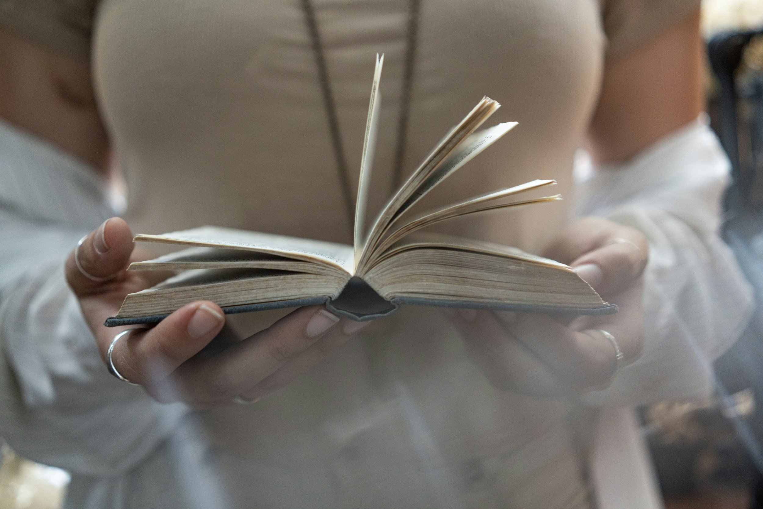 A person holding an open book with the person's torso and hands visible. The background is blurred.