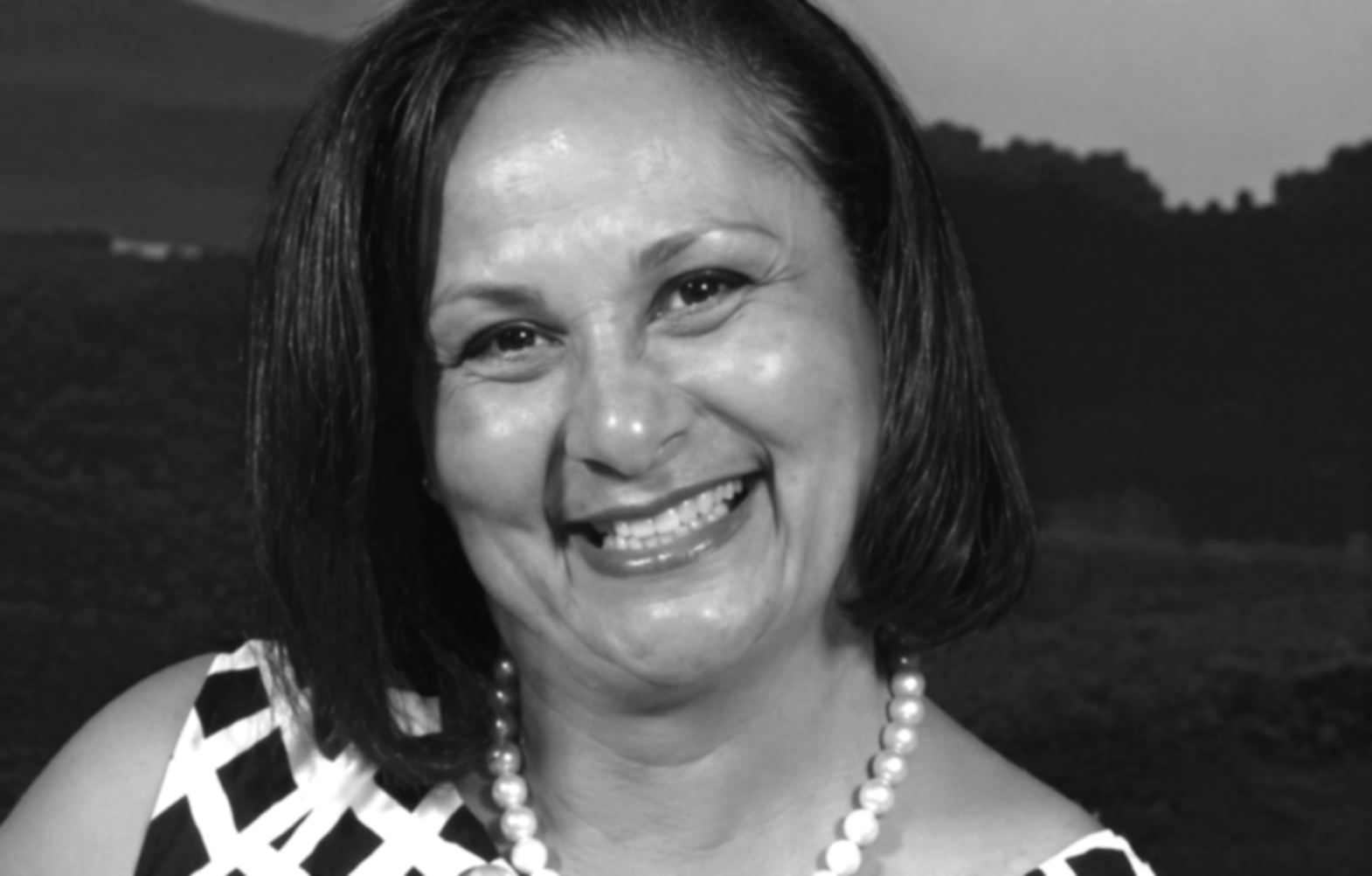 A woman smiling, wearing a sleeveless patterned dress and a pearl necklace, with short dark hair, outdoors with hills and trees in the background.