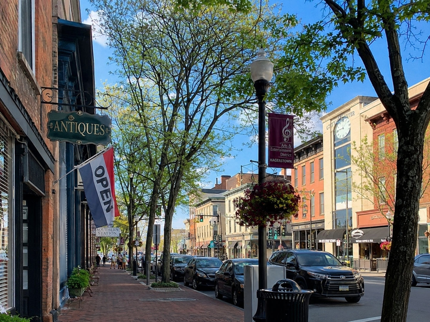 Downtown street scene with shops, trees, parked cars, hanging flower basket, lamp post, and signs including an antiques shop, open sign, and arts & entertainment banner on a sunny day.
