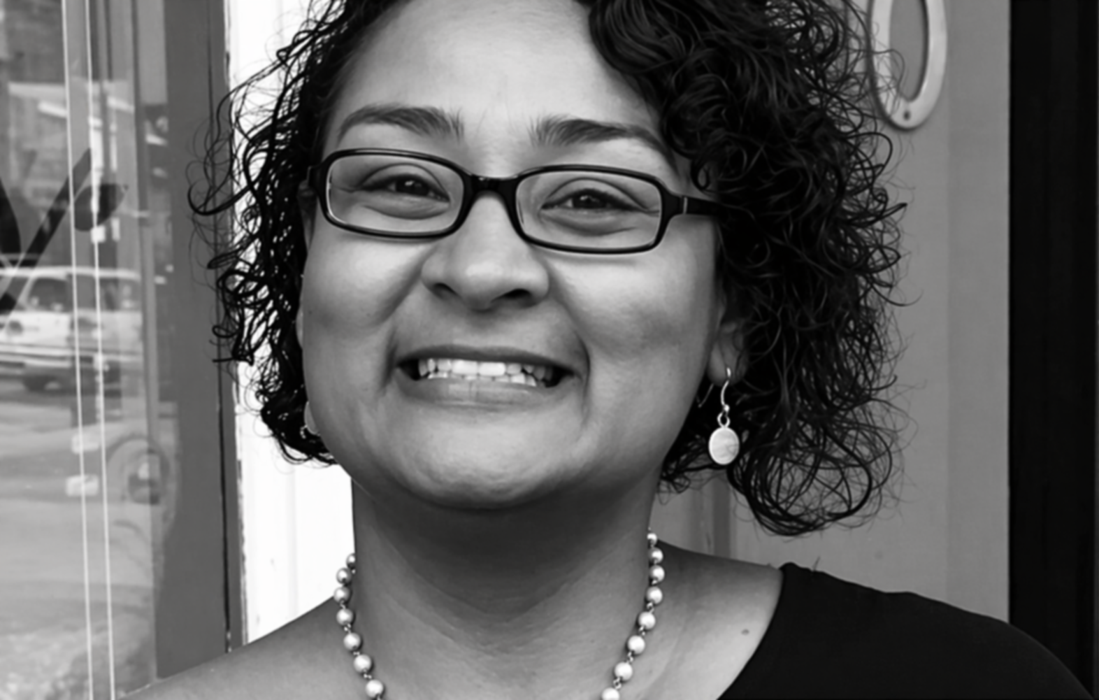 A black and white photograph of a woman with curly hair, glasses, earrings, a necklace, smiling, outdoors with a reflection of a street and cars in the background.