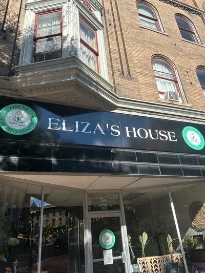 The storefront sign reads "Eliza's House" with a black background, green circular logos on either side, and the building above features a bay window with curtains and brick exterior.