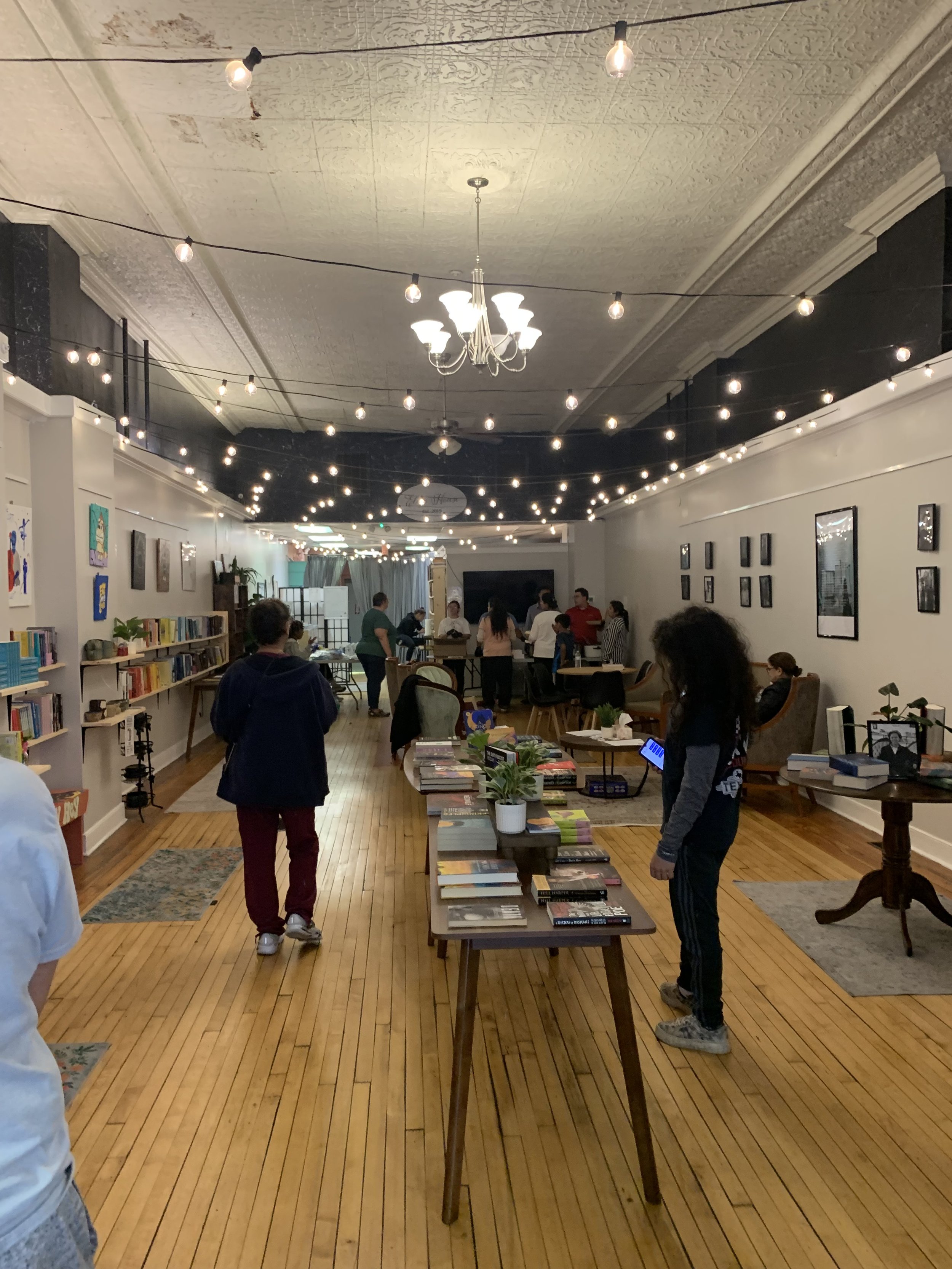 Interior of a bookstore or library with bookshelves, tables filled with books, and patrons reading or browsing. The ceiling has string lights and a chandelier, and the floor is wooden.
