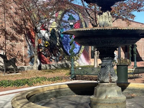A decorative water fountain in the foreground with an ornate design, in front of a colorful mural on a brick wall, featuring artistic figures and abstract shapes, with benches and trees nearby on a sunny day.