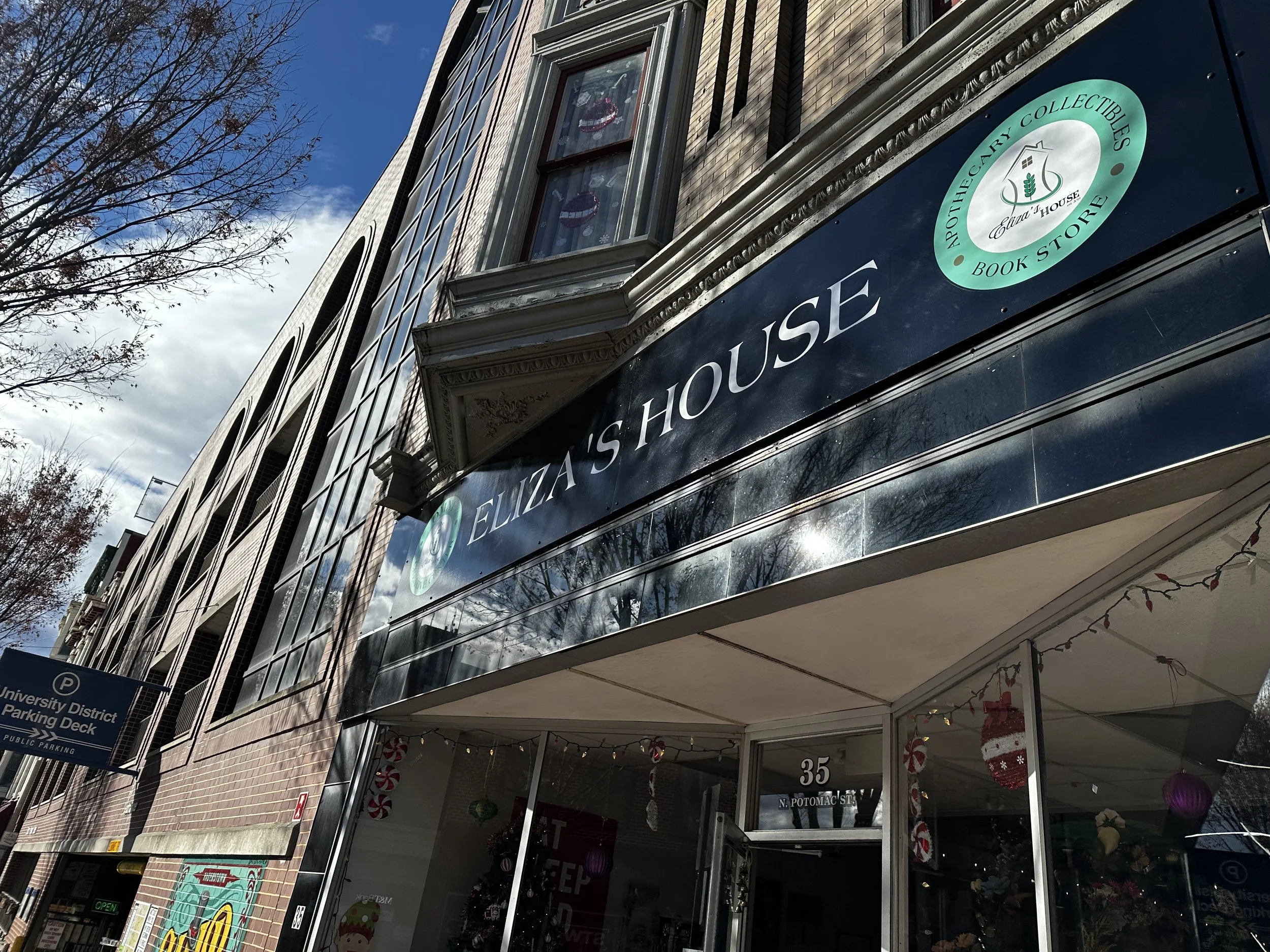 The exterior of a building with a sign that reads 'Eliza's House' and 'Resale & Collectibles Book Store,' with holiday decorations visible in the window and some trees with bare branches against a partly cloudy sky.