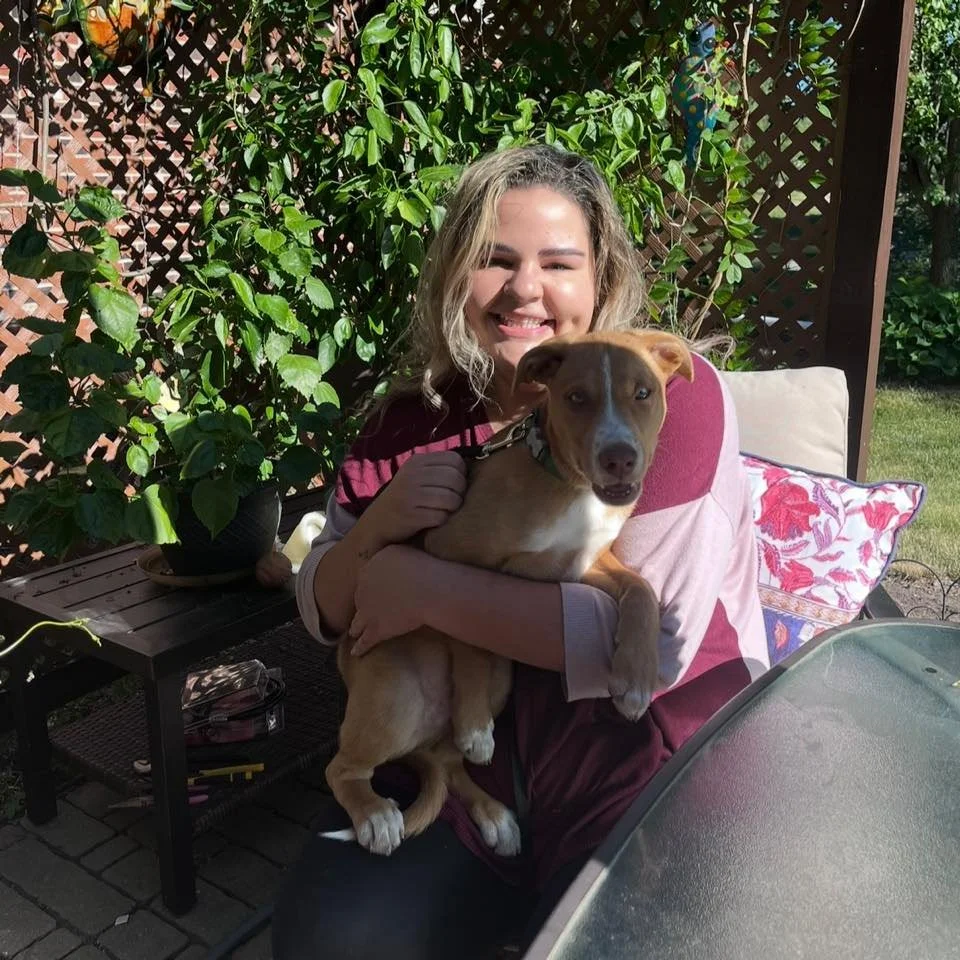 A young woman with curly blonde hair smiling and holding a brown and white dog outside in a garden area. The woman is sitting on a patio with lush greenery and a wooden lattice in the background, and there are decorative pillows on a chair beside her.