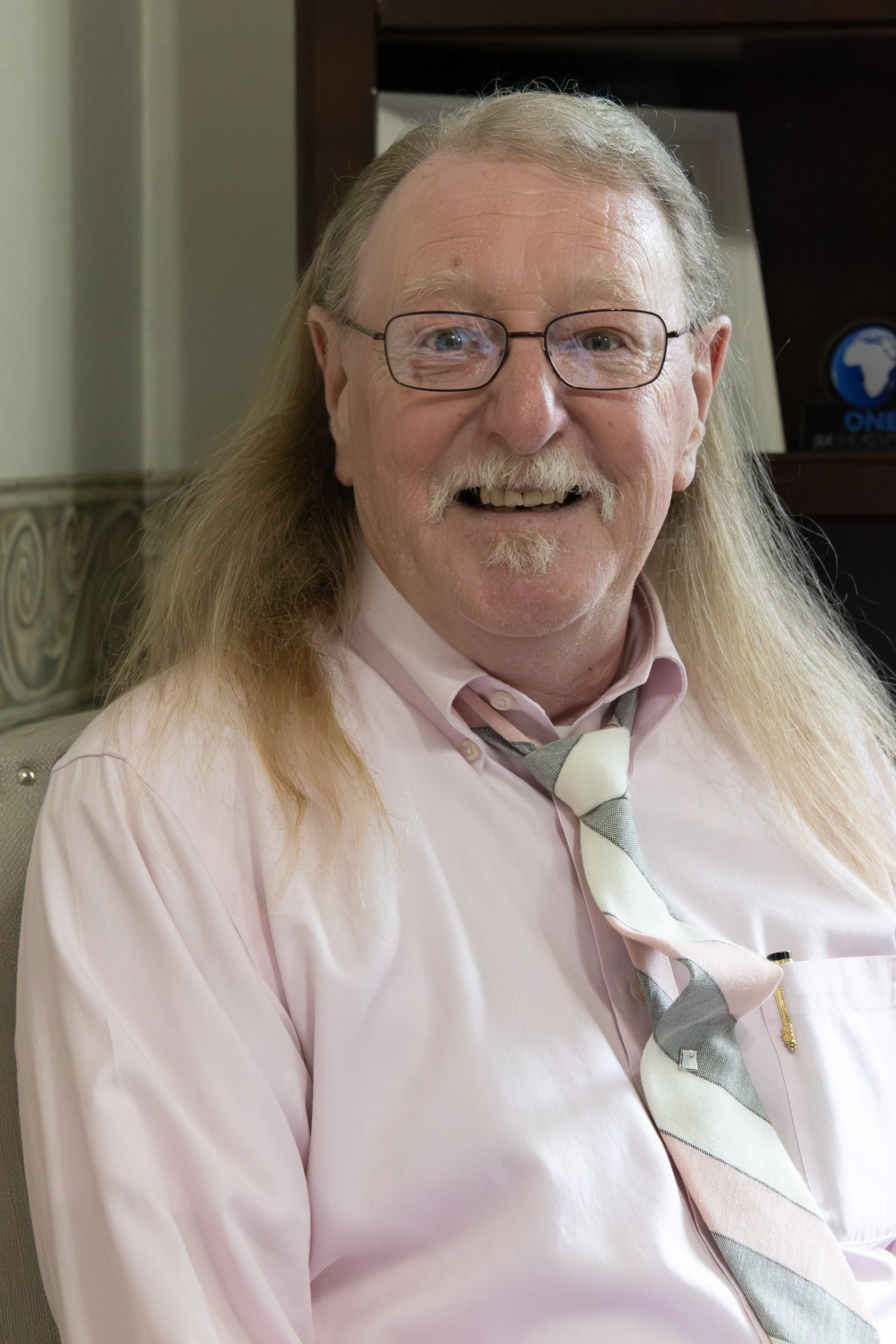 Portrait of Sandy Smith, a smiling elderly man with long gray hair, glasses, and a mustache, wearing a pink shirt and a striped tie, sitting indoors with a bookshelf and a decorative wall in the background.