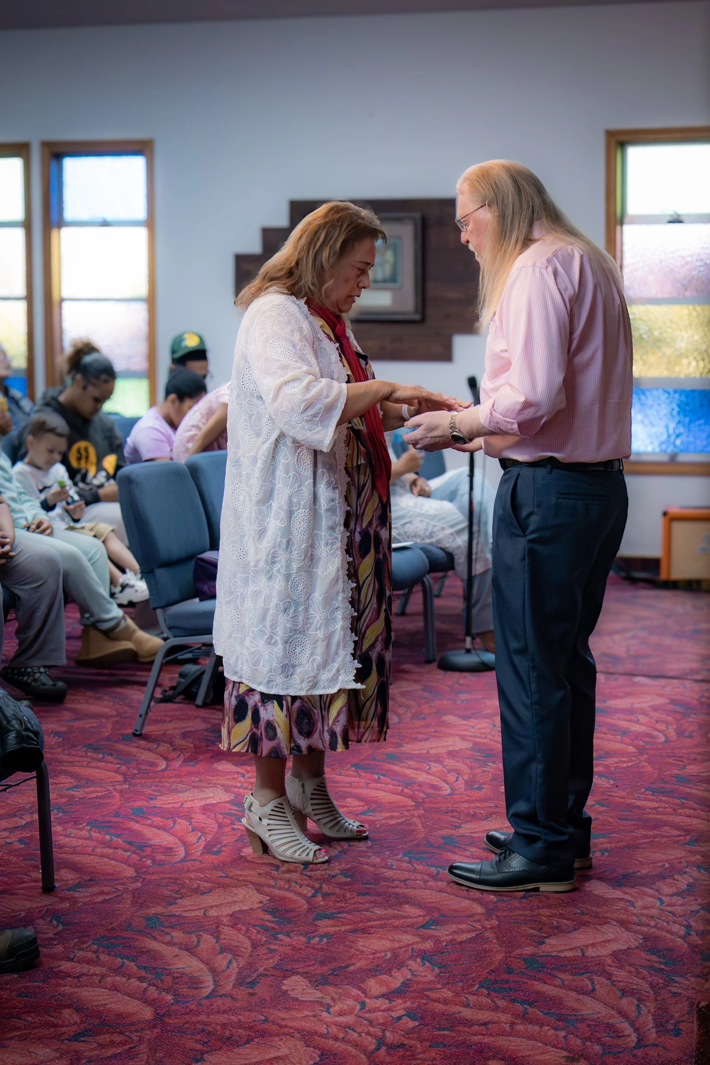 A woman in a white lace dress and beige heels receiving prayer from a man in a pink shirt and dark pants during a ceremony in a room with red patterned carpet and seated congregation.