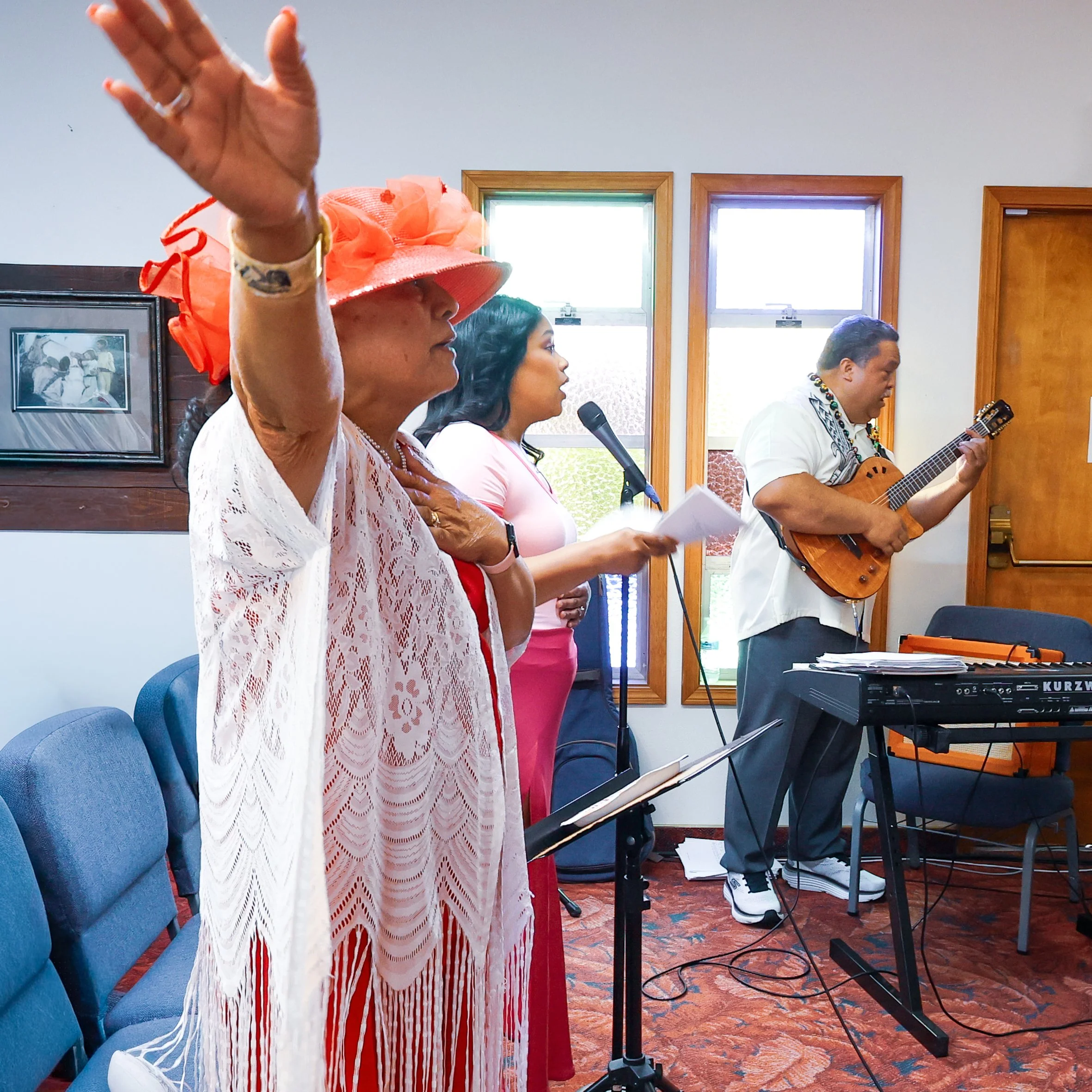 A group of people singing and playing guitar during a musical performance or worship service, with sunlight coming through windows.