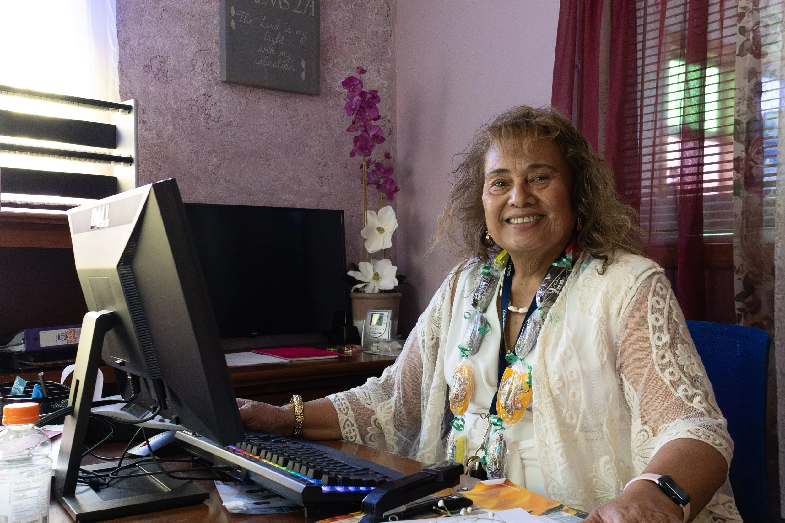 A woman sitting at a desk in an office, smiling at the camera, with a computer monitor, keyboard, and various office supplies visible. The background includes purple textured wall art, flowers on a side table, and pink curtains.
