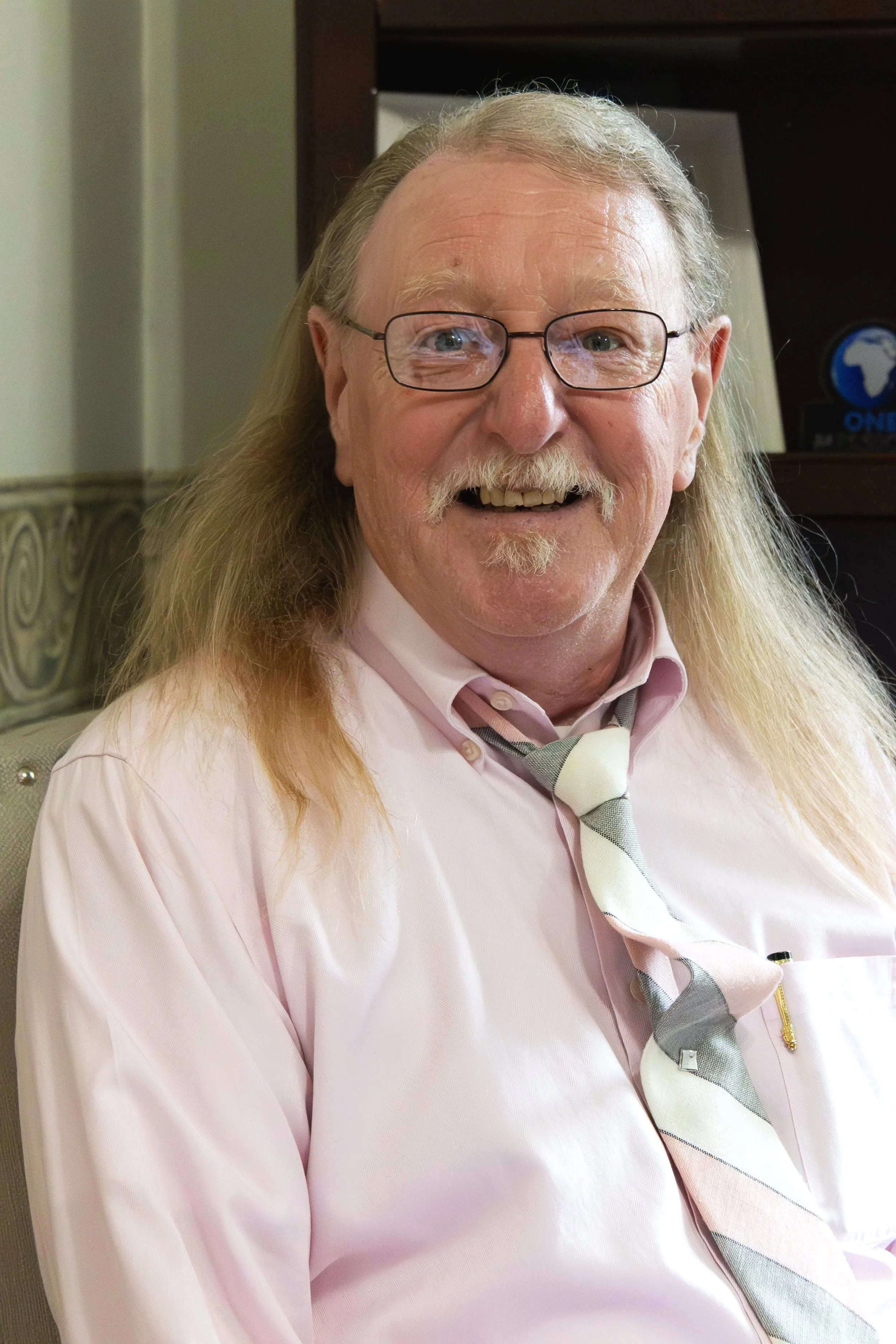 A smiling elderly man with long gray hair, glasses, and a mustache wearing a pink shirt and a striped tie.