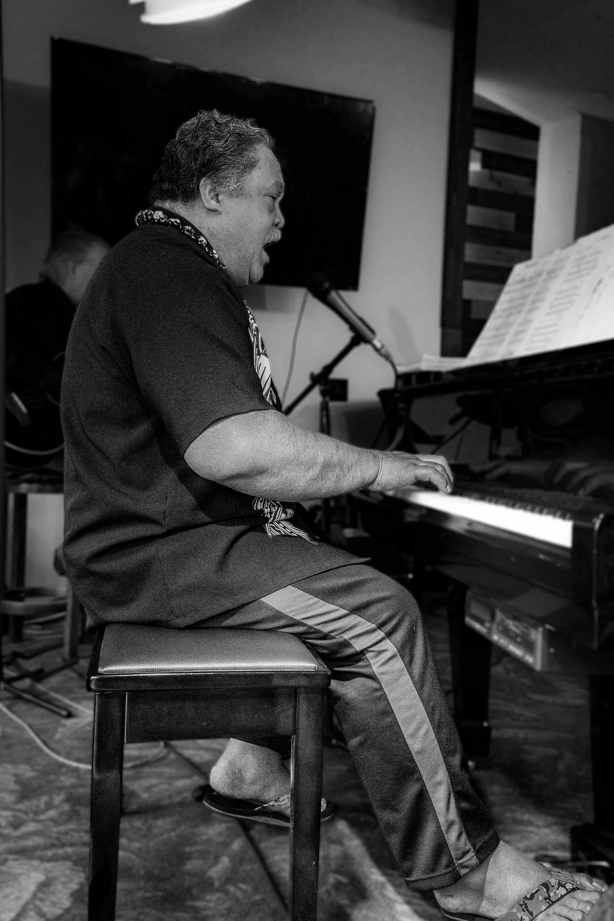 A man playing the piano on a stool, singing into a microphone, with music sheets on the piano, in a room with a television and wooden wall decor in the background.