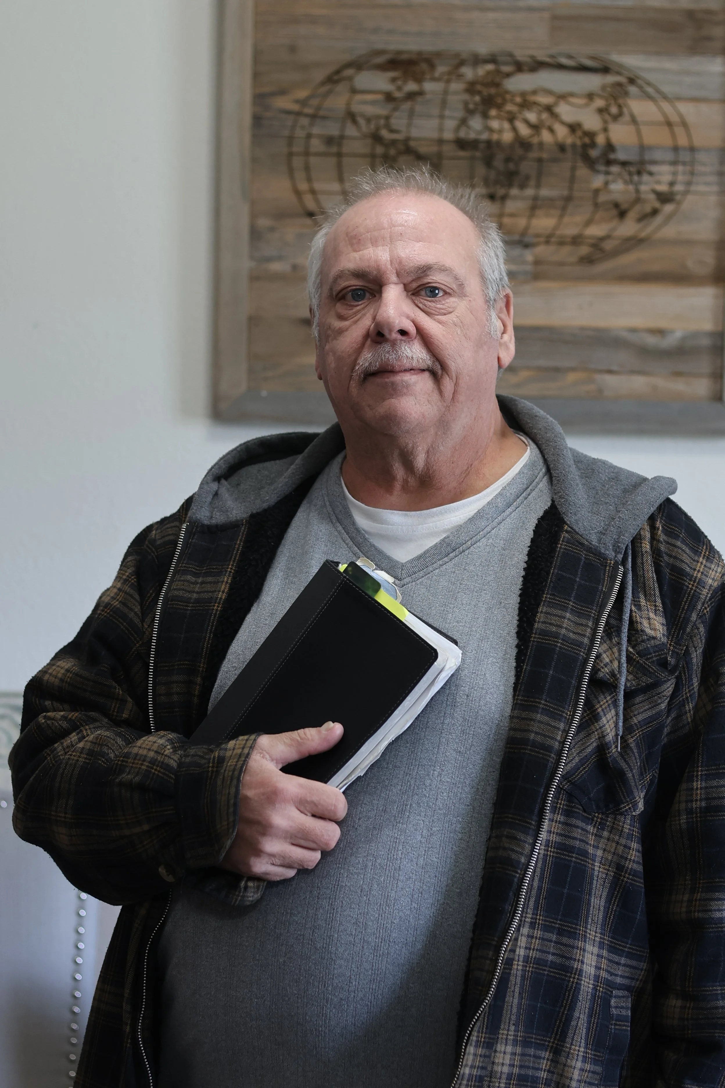 A middle-aged man with gray hair and a mustache, wearing a gray shirt and a brown plaid jacket, holding a clipboard, standing in front of a wall with a wooden world map.