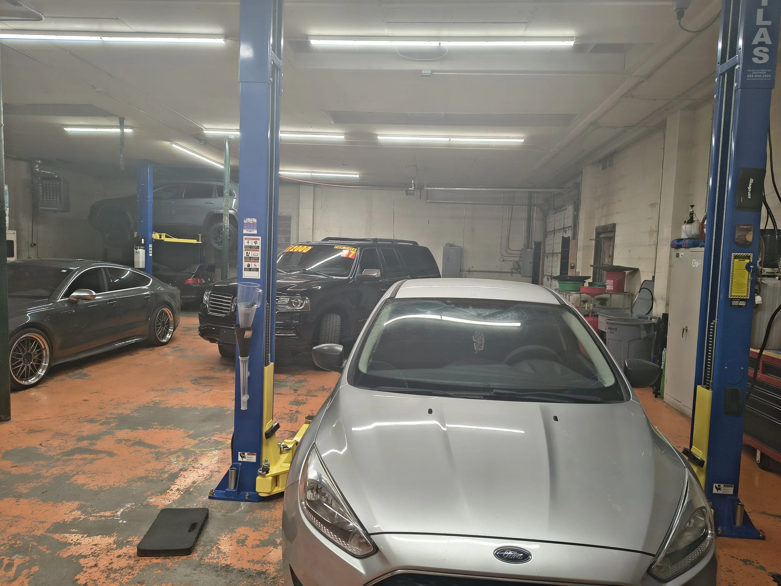 An indoor car repair shop with multiple cars, including a silver Ford in the foreground and a black SUV on a lift in the background, with bright fluorescent lighting and various tools and equipment.