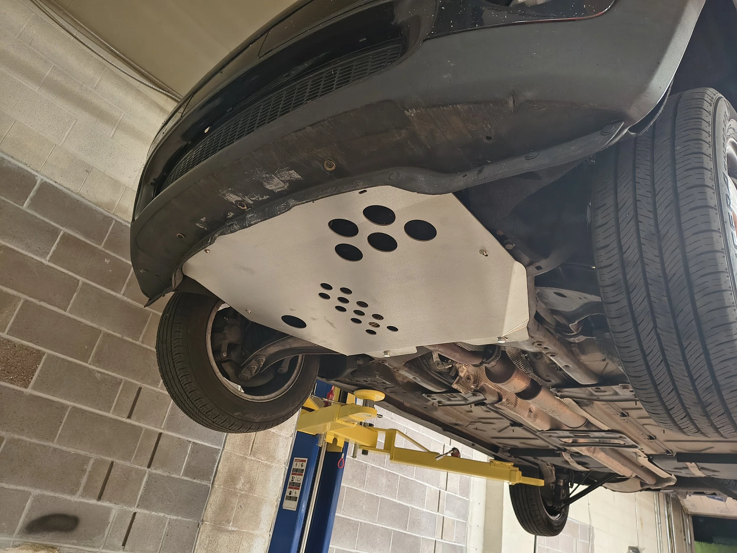 Underneath view of a black car on a hydraulic lift in a garage, showing the underside and rear tire.