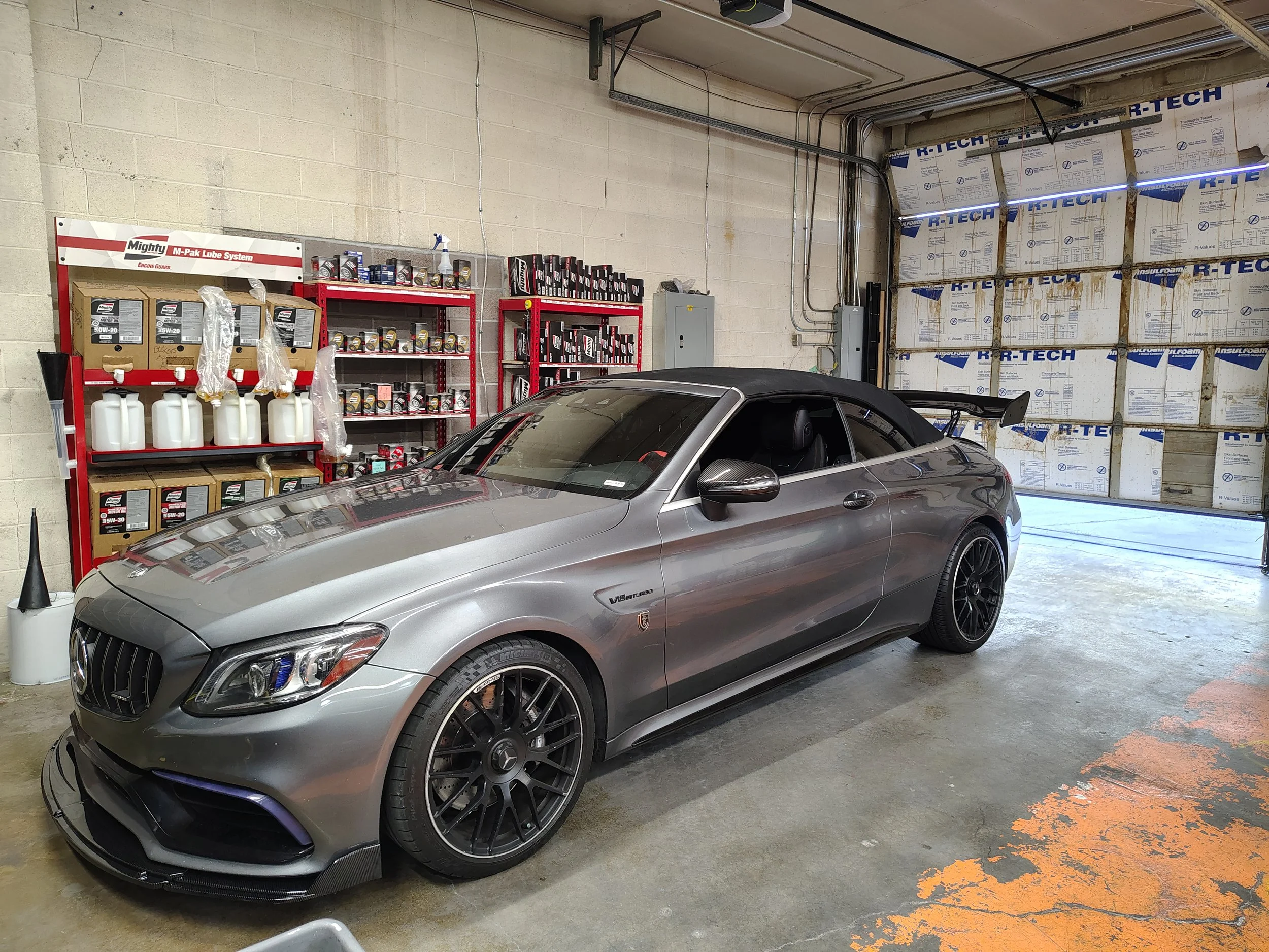 A silver Mercedes-Benz sports car parked inside a garage. The garage has exposed concrete walls, shelves with car maintenance supplies, and insulation panels on one wall.