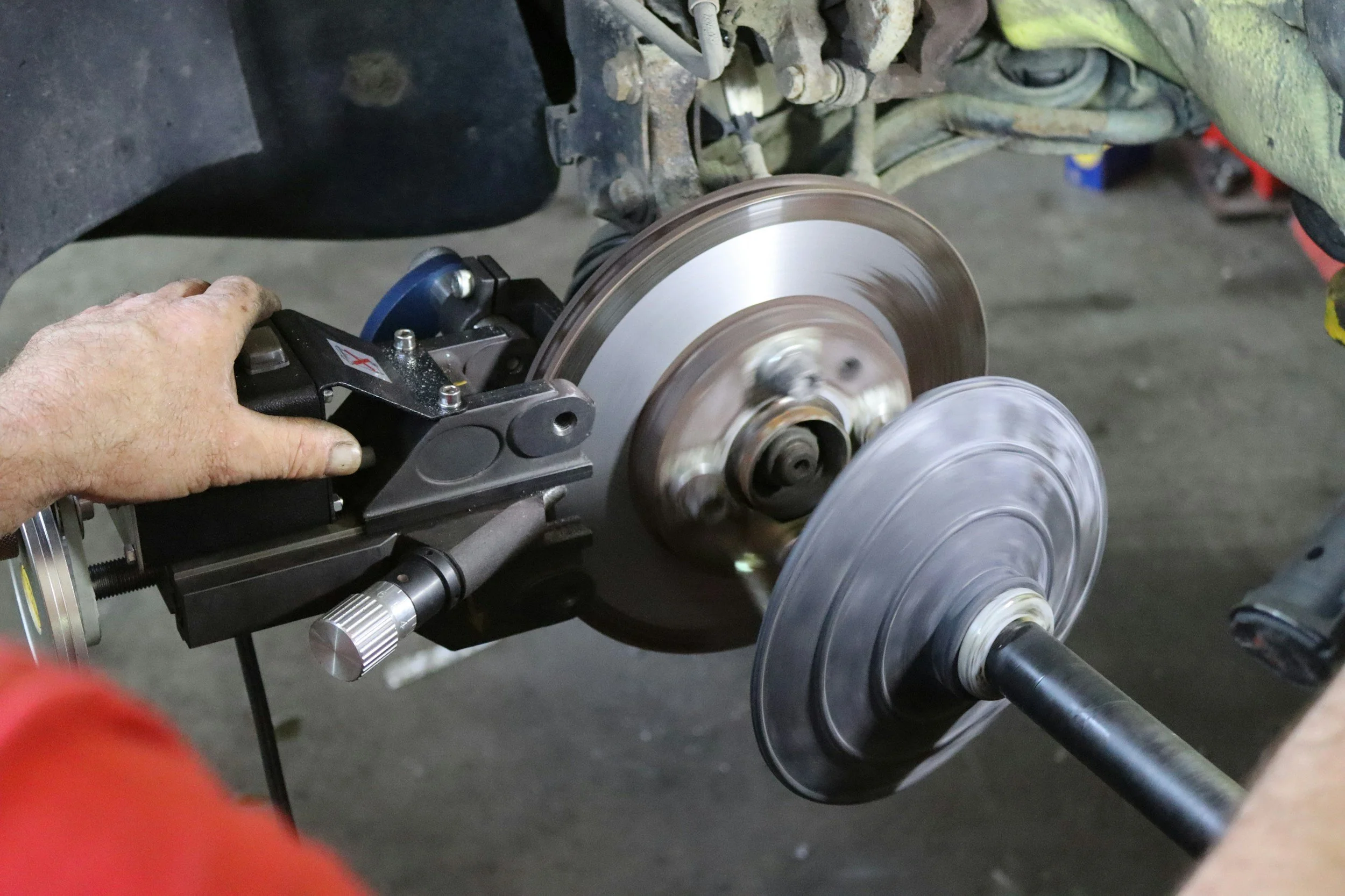 A person wearing red gloves uses a power tool to cut a brake disc on a vehicle's wheel assembly.