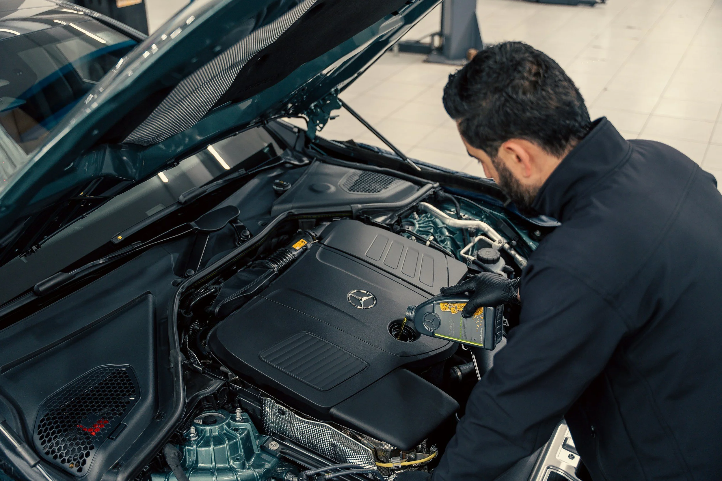 A man with dark, curly hair and a beard inspecting the engine of a dark green Mercedes-Benz with the hood open inside a showroom or garage.