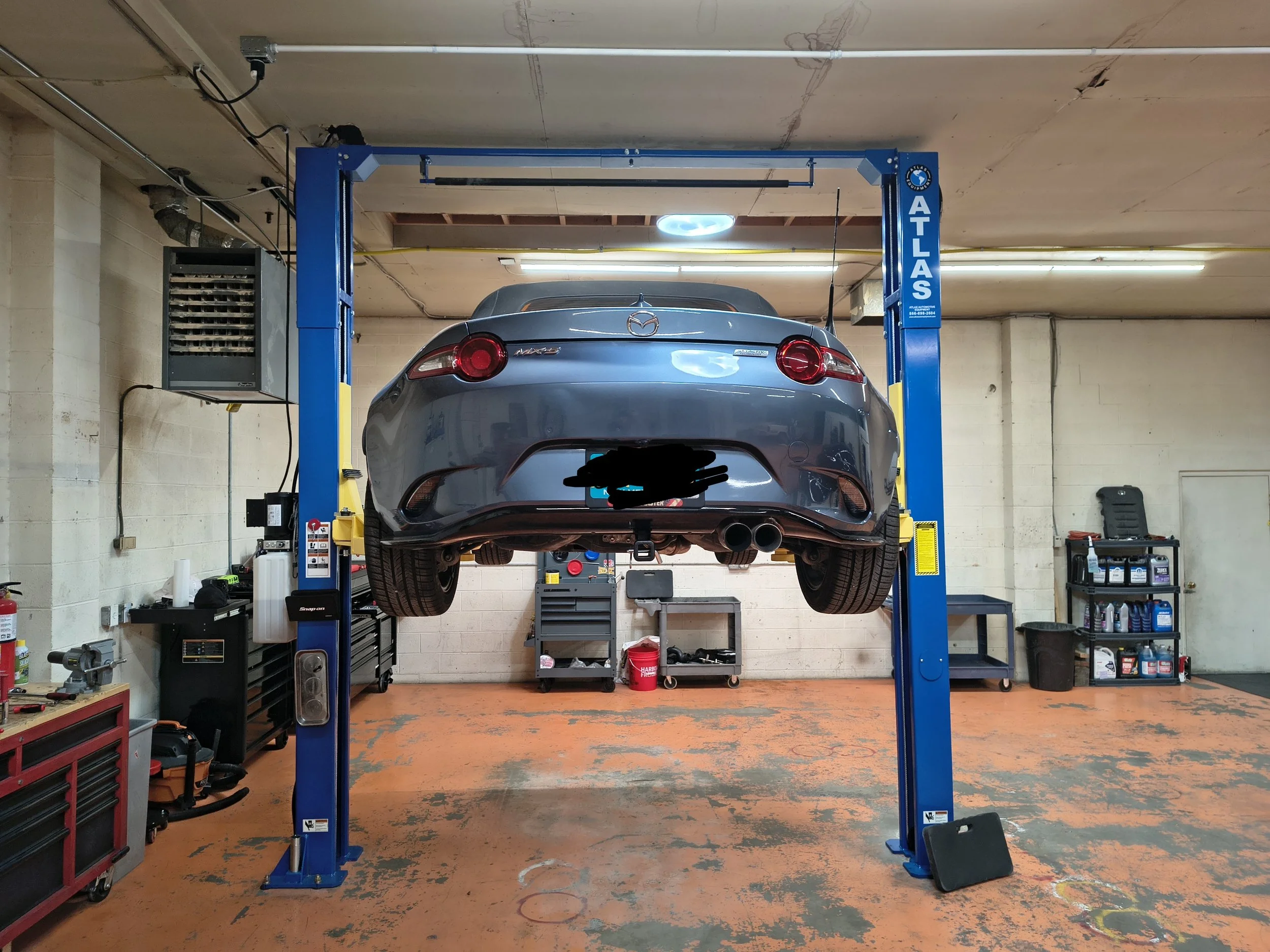 A gray Mazda MX-6 car is elevated on a blue hydraulic lift inside an auto repair shop. The workshop has beige cinder block walls, various tools, and equipment on shelves and workbenches. The concrete floor shows orange paint with some wear and stains.