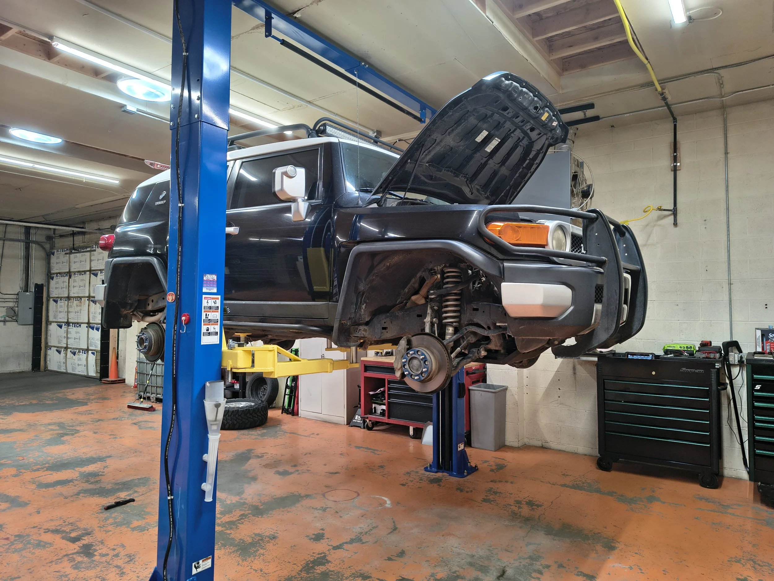 A black SUV is lifted on a blue vehicle lift in a garage, with its hood open for maintenance or repair. The garage has an industrial look with a concrete floor, exposed ceiling beams, fluorescent lighting, and various tools and equipment around.