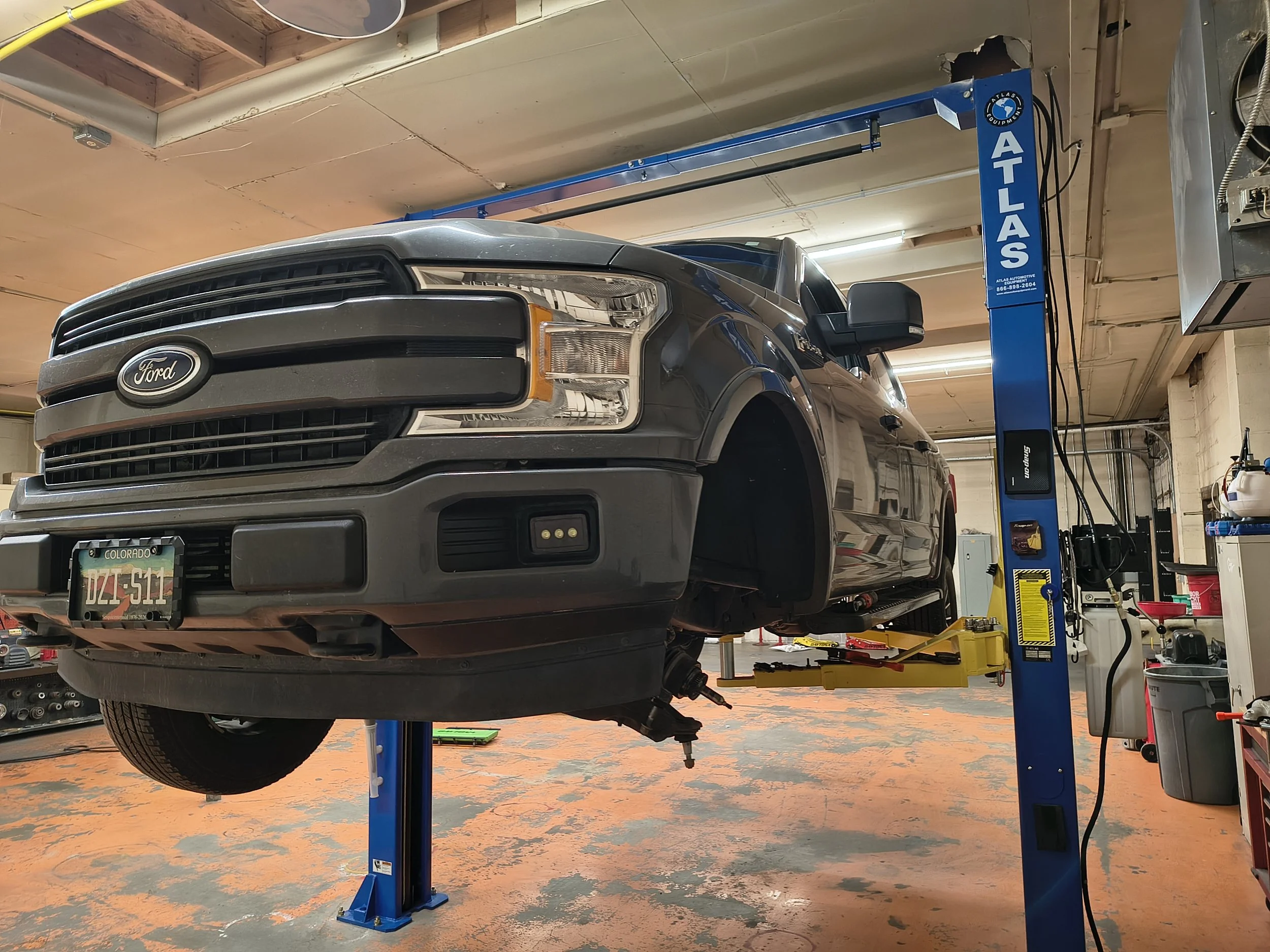 Front view of a dark gray Ford pickup truck lifted on a blue hydraulic vehicle lift inside an auto repair garage. The garage has a beige ceiling, orange and gray concrete floor, and various tools and equipment in the background.