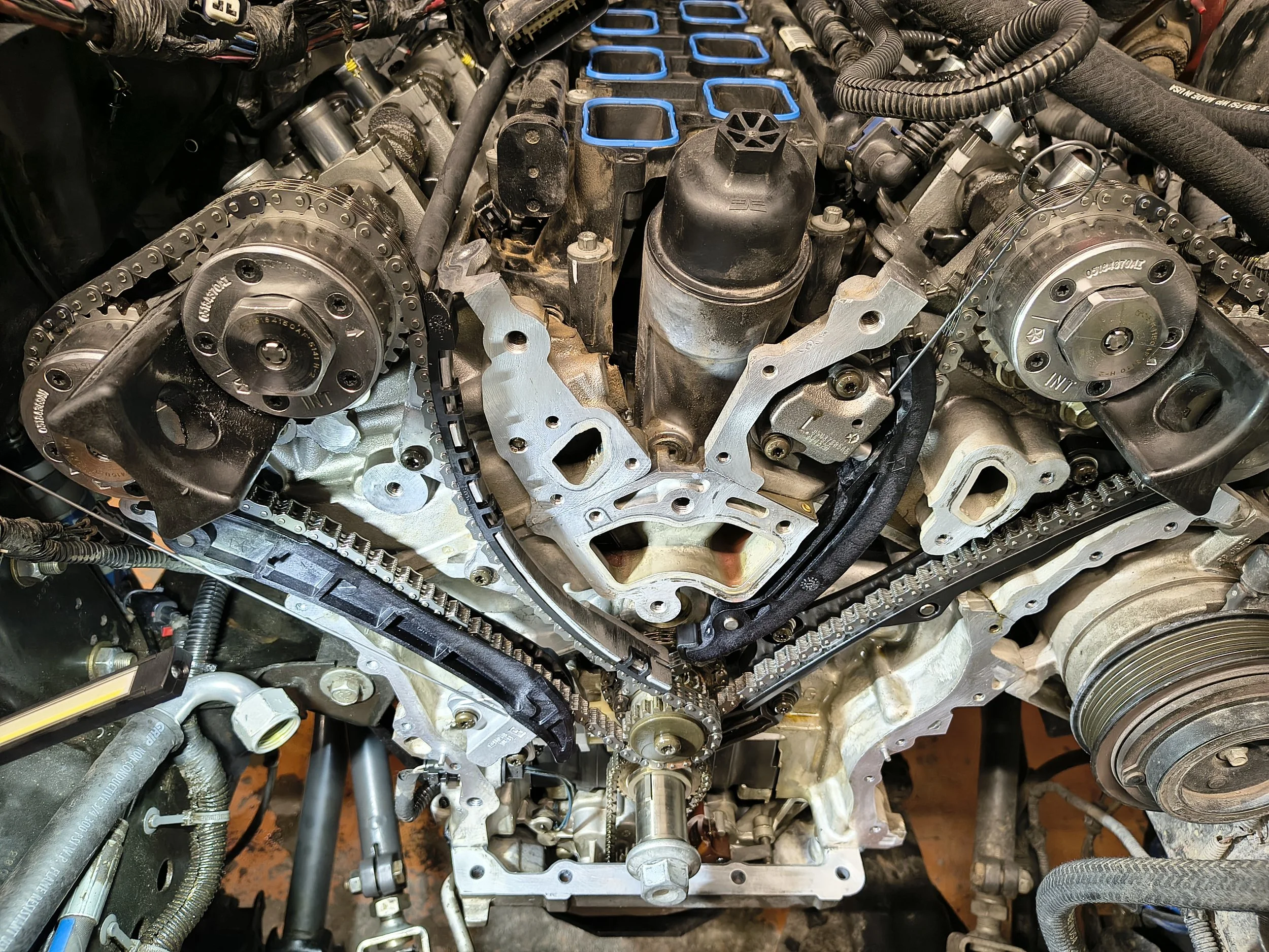 Close-up view of a car engine with visible timing chain, pulleys, and other engine parts, showing an engine in the process of repair or assembly.