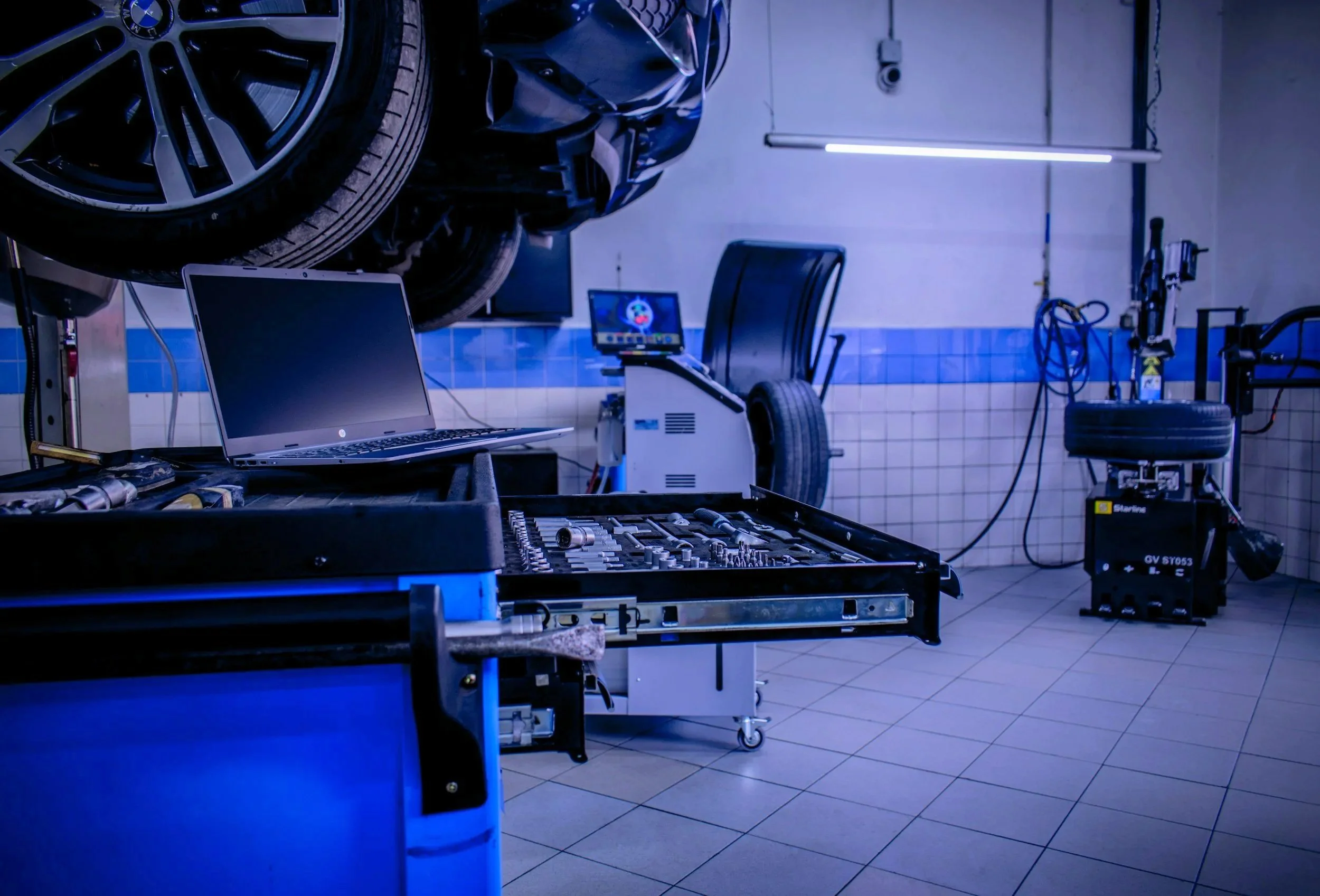 Automotive workshop with a car on a lift, tools, a laptop, and diagnostic equipment in a garage with tiled flooring and fluorescent lighting.