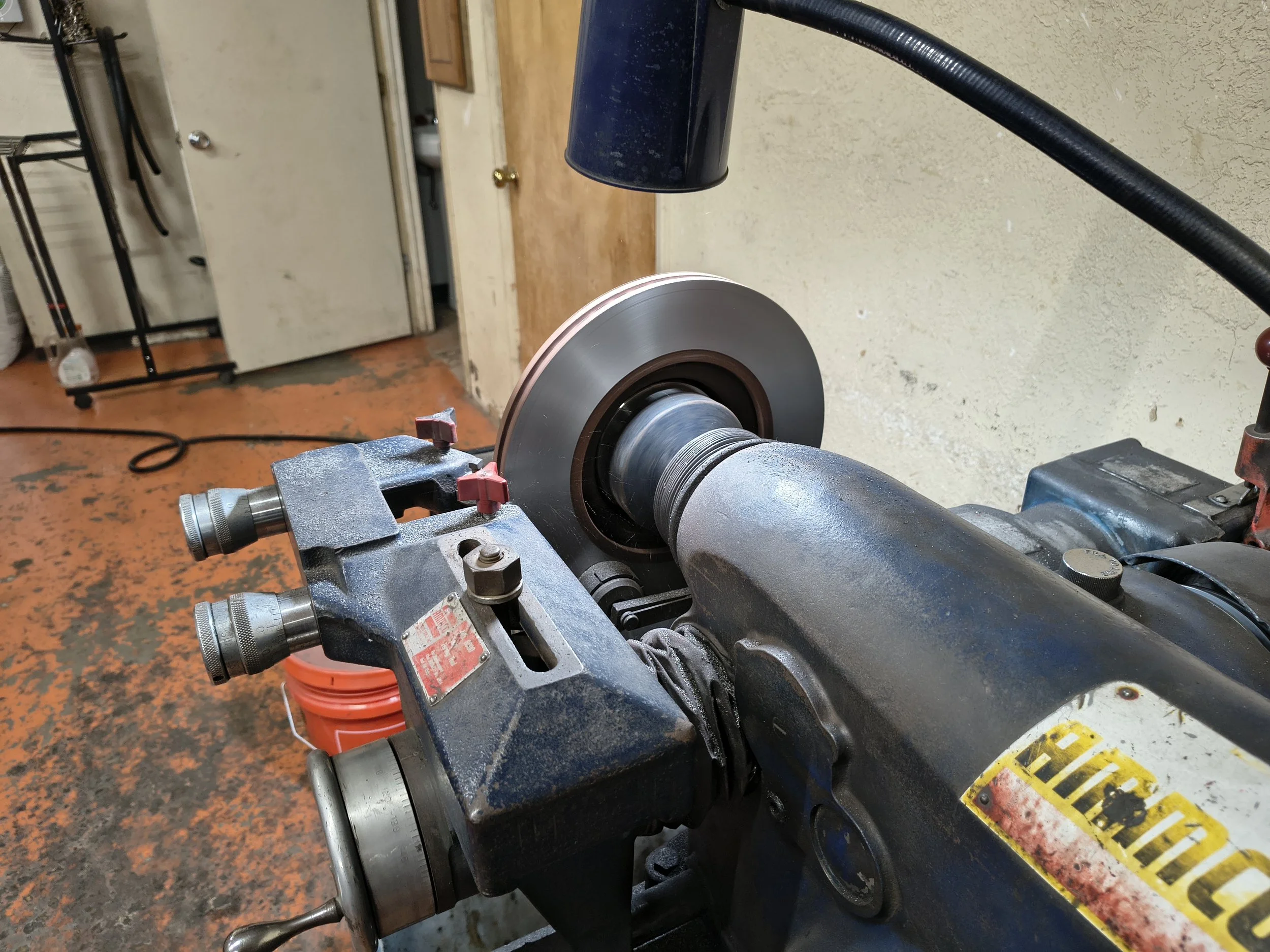 Workshop environment with a rotary tool machine in focus, a red bucket underneath, and a workbench with tools and a spray bottle in the background.