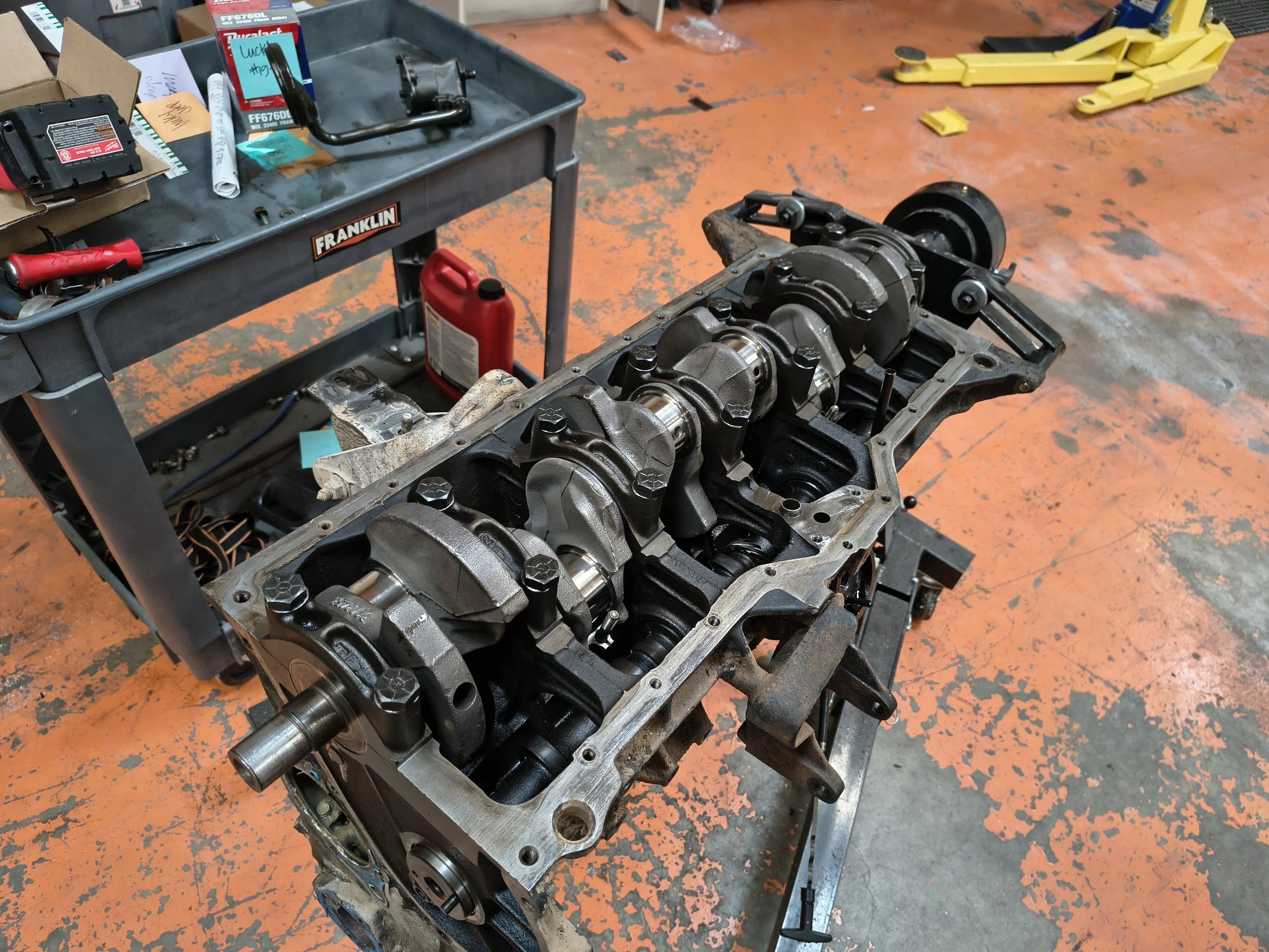 A partially assembled engine block with pistons visible, placed on a stand in a workshop. A workbench with tools, a red oil container, and yellow equipment are in the background, along with an orange industrial floor.