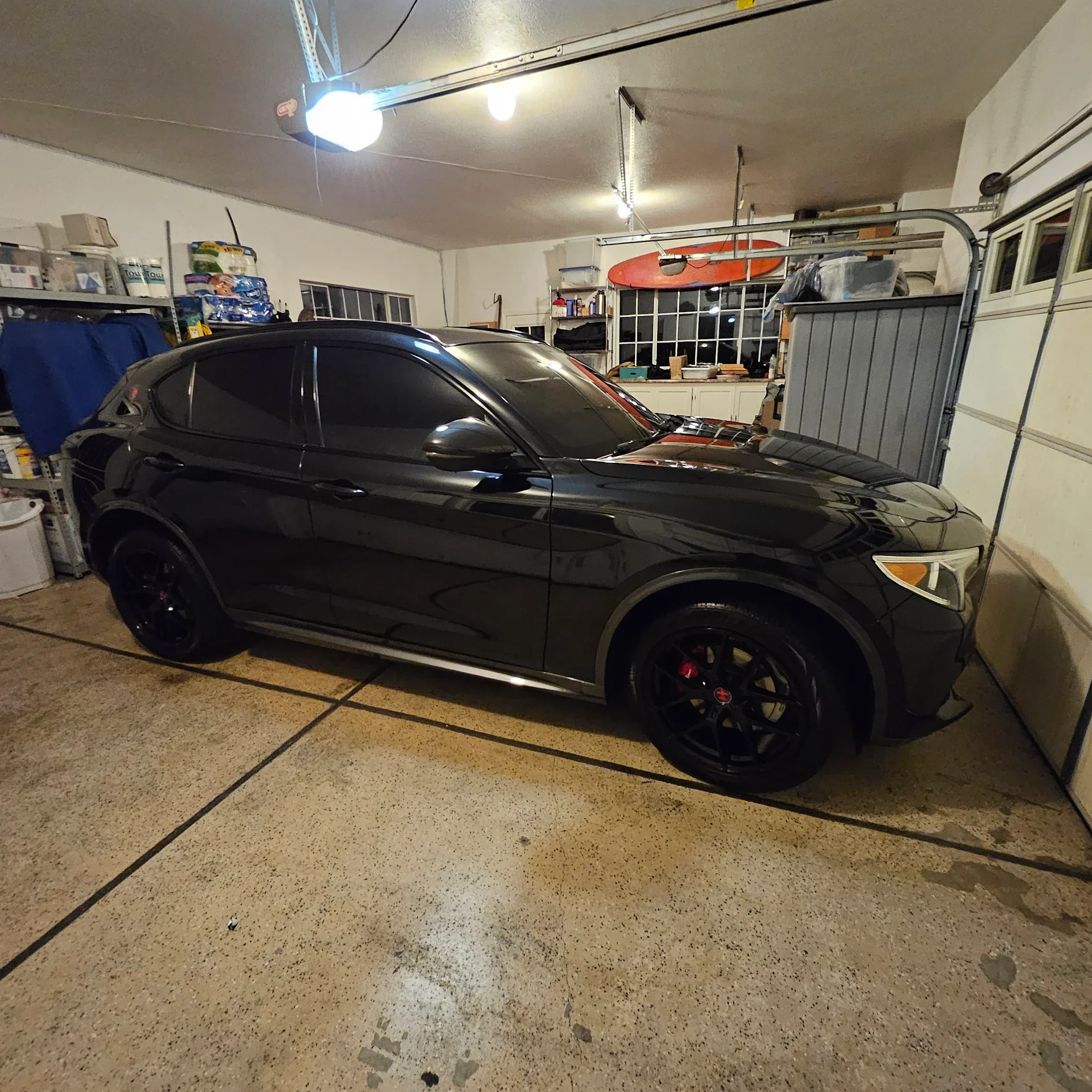 A black sports car parked in a garage with tools, supplies, and a surfboard on the wall.