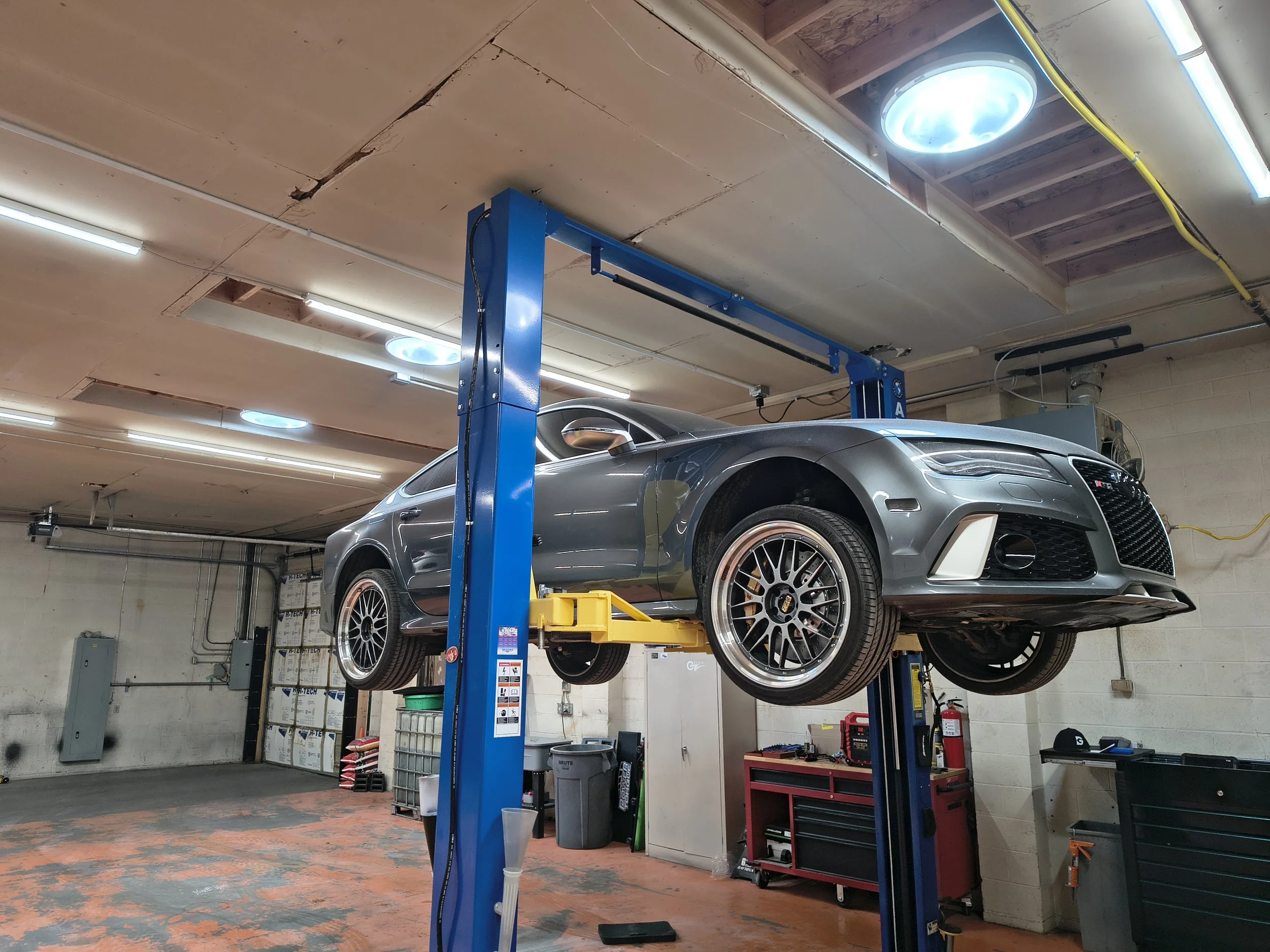 A silver sports car raised on a blue hydraulic lift in an auto repair garage.