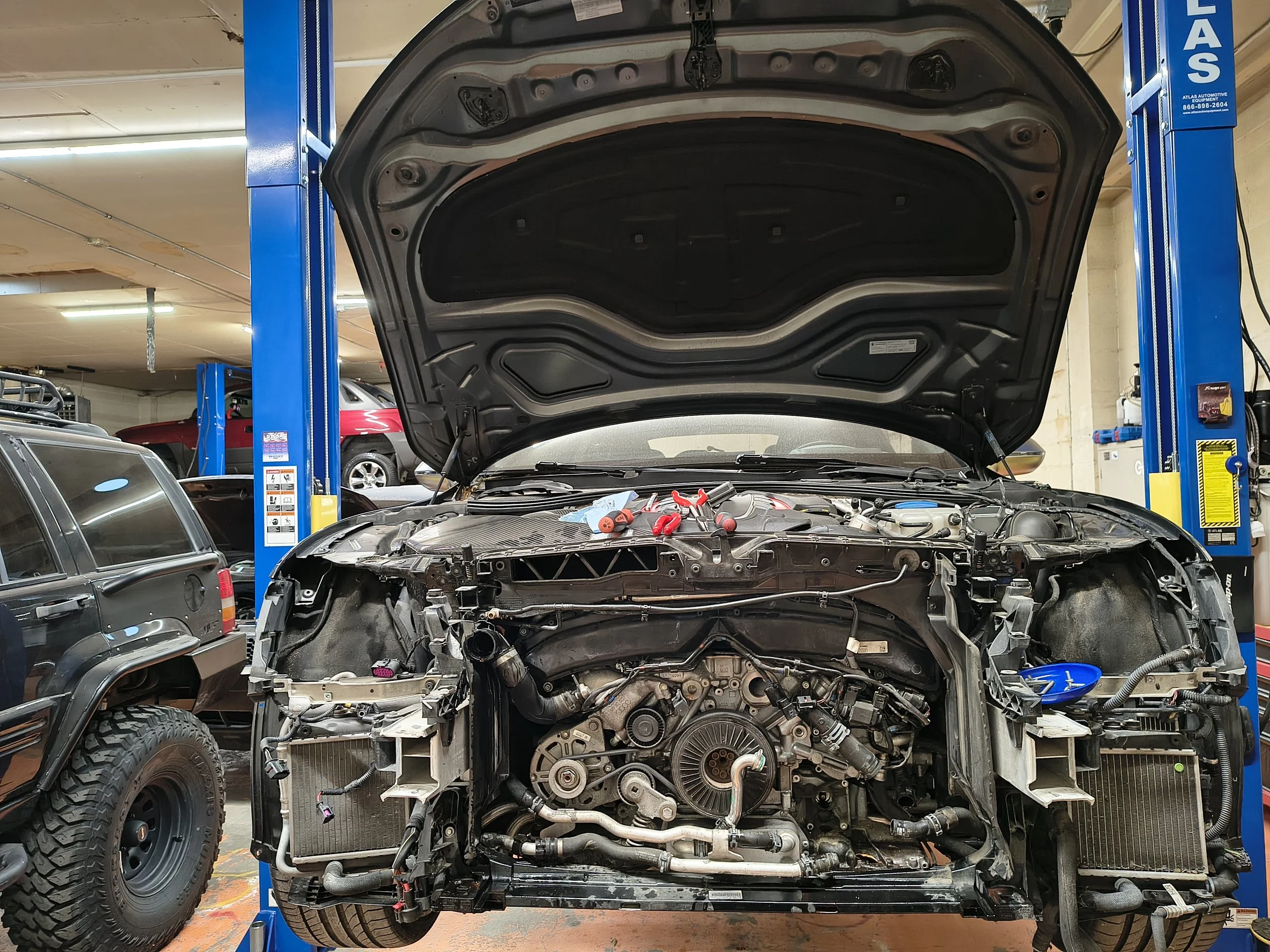A car with its hood open on a lift at an auto repair shop, exposing the engine and internal components of the vehicle.
